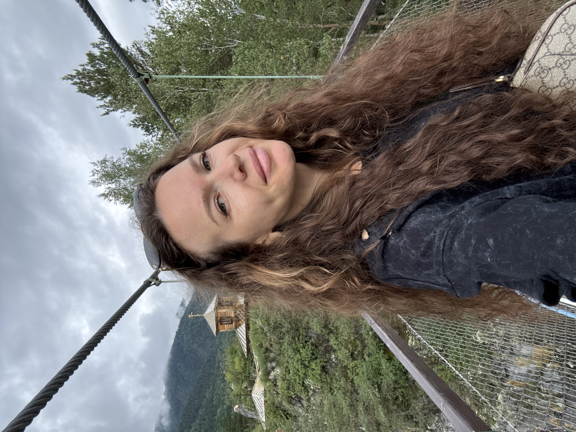 A woman with long, wavy brown hair takes a selfie on a suspension bridge. She is wearing a black hoodie and sunglasses are perched on top of her head. In the background, a wooden church with a golden cross on its roof sits atop a hill covered in green foliage. Mountains and trees form the backdrop under a cloudy sky. The scene is likely located near the Chemal, Russia, area, possibly at the Patmos Island monastery, which is accessible by a suspension bridge.