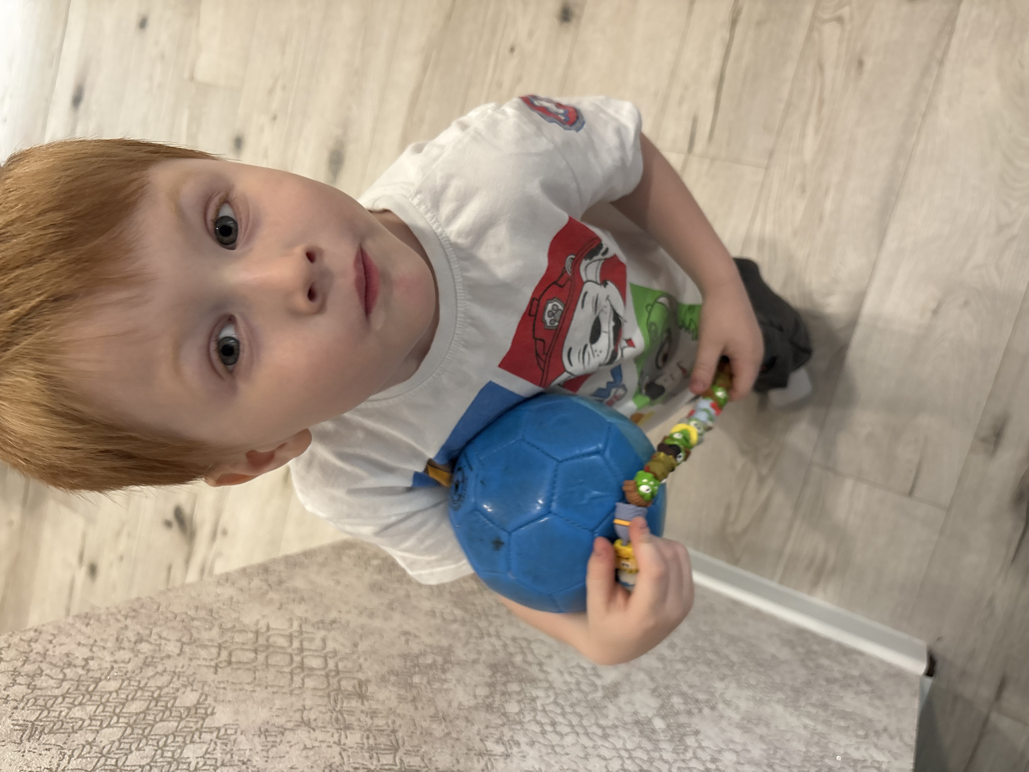 A young, fair-skinned child with red hair looks up at the camera. The child wears a white t-shirt with a graphic cartoon design. The child holds a blue soccer ball in one hand and a colorful string toy in the other. The background shows a light-colored wood-look floor. The bottom left corner shows a countertop or table with a neutral, textured pattern. The location is unknown.