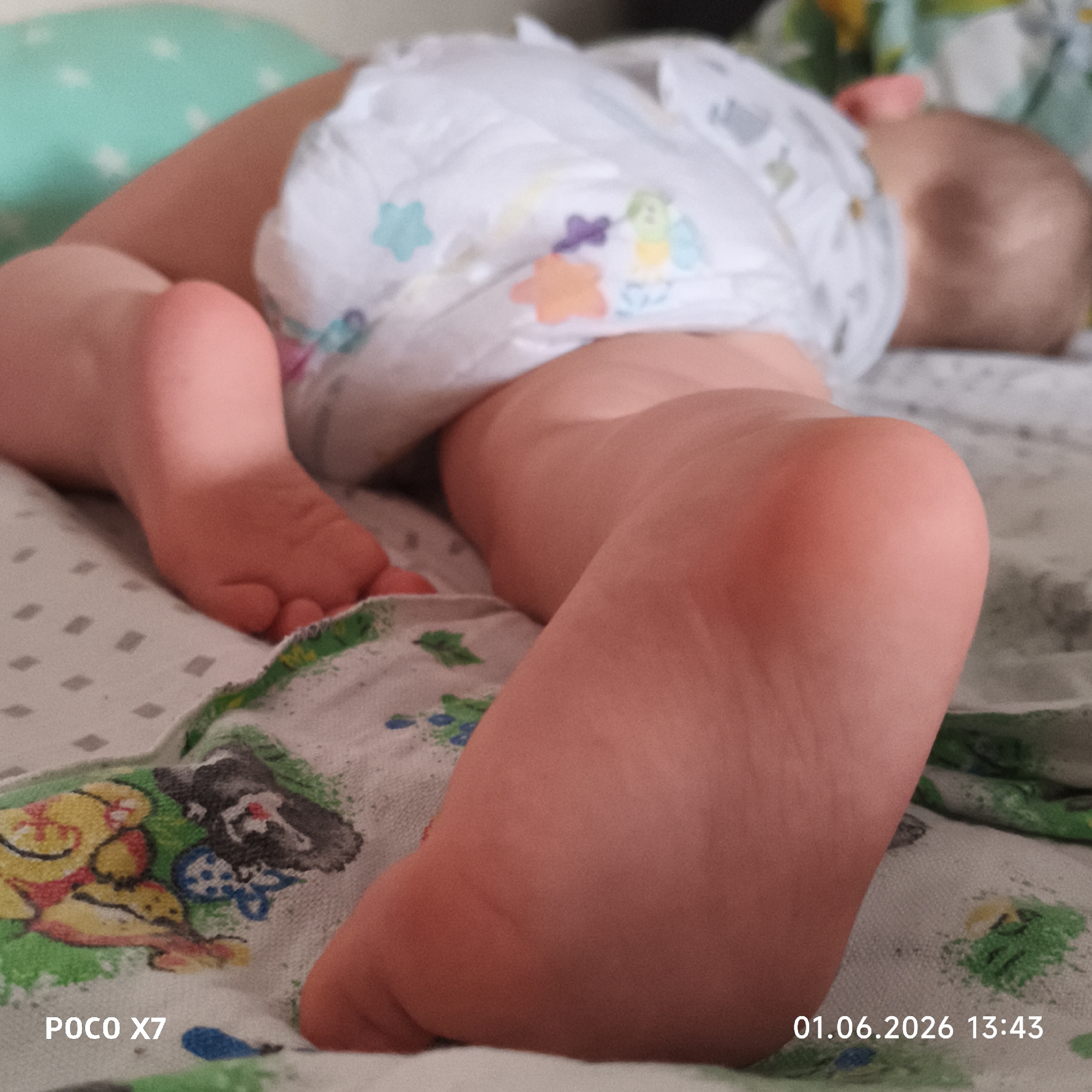 A close-up photograph shows a baby lying prone on a patterned bed sheet. The baby is wearing a diaper with cartoon-like decorations. The baby's feet are prominently displayed in the foreground. A portion of the baby's head is visible in the background. The photograph was taken on "01.06.2026 13:43" with a "POCO X7" camera. The location of the photograph is unknown; it is not possible to determine if it was taken in Chaikovsky, Russia from this image alone.