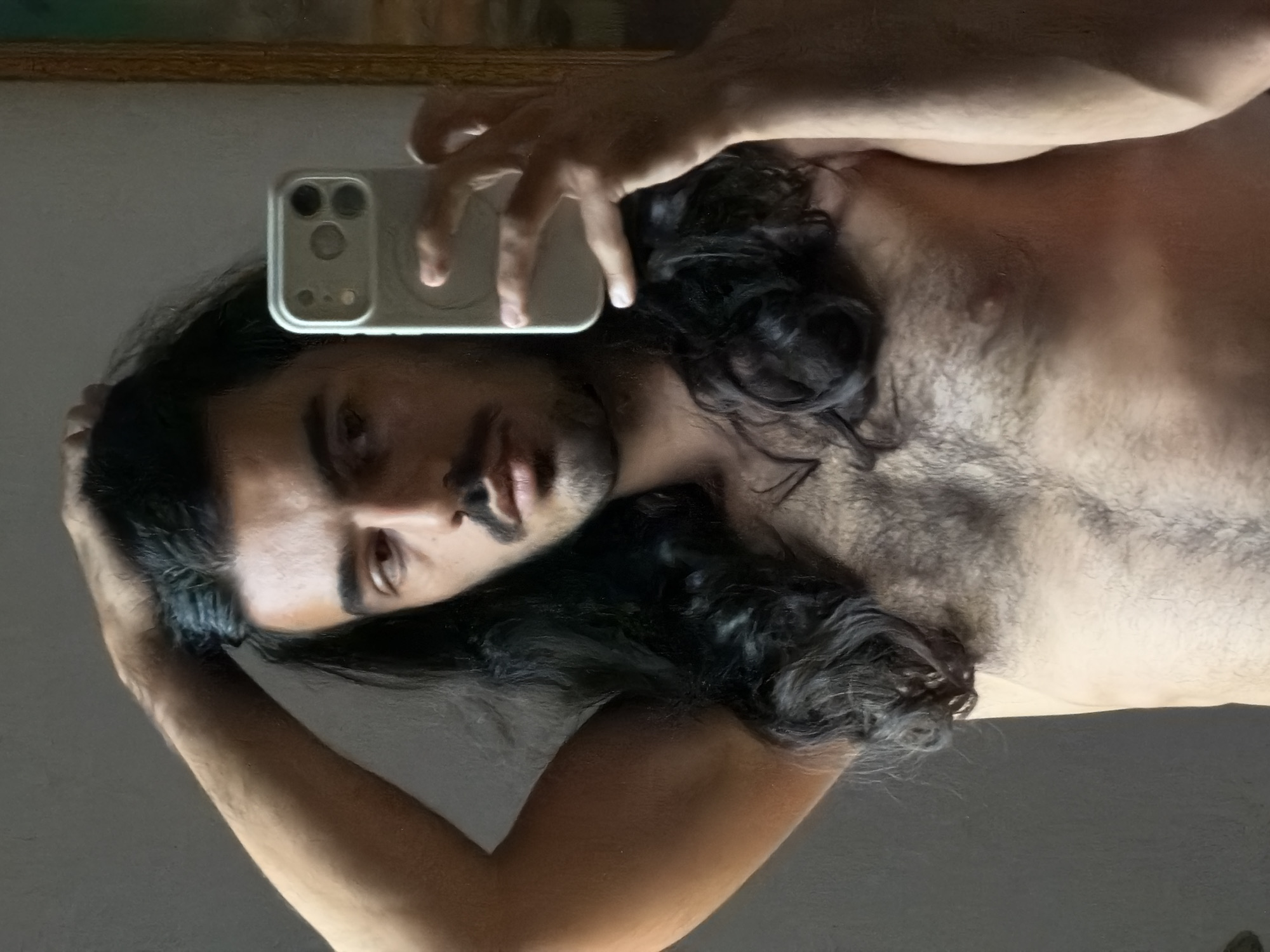 A man is taking a selfie in a mirror. He has dark hair, a mustache, and a hairy chest. He is holding a white smartphone with multiple camera lenses. The location is not discernible from the image.