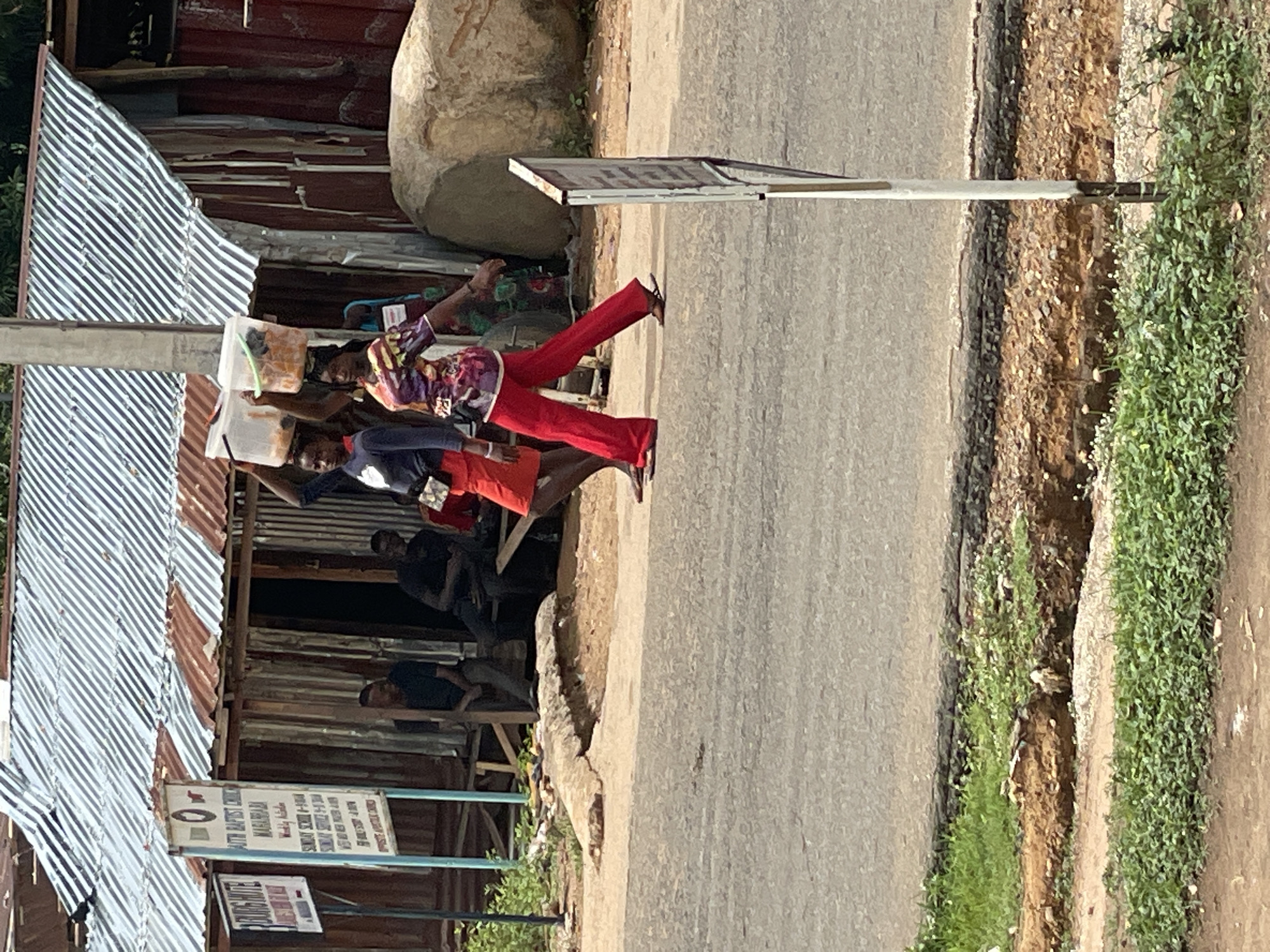 The image shows a street scene in Mararaba, Nigeria. Several people are visible. One woman is wearing red pants and a patterned top, walking along the side of the road. There are other people sitting under a corrugated metal roof structure near a sign. The sign indicates directions to a "South Baptist Church Mararaba," with times for Sunday School and Sunday service. A street and a grassy area are also visible. A large rock sits above the metal structure.