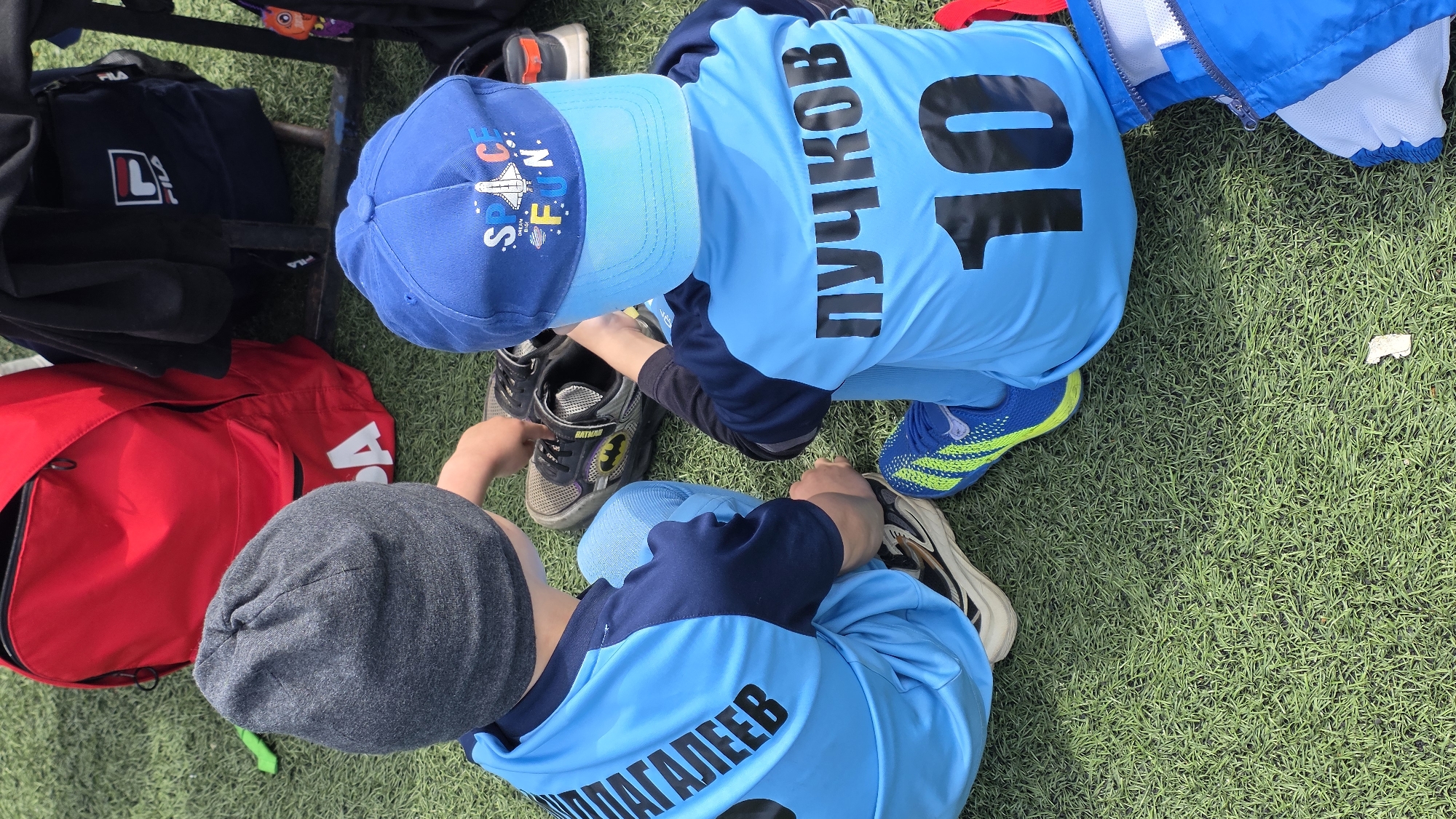Two children sit on a green surface. One child wears a blue cap with "SPACE FUN" printed on it and a light blue jersey with the number "10" and the name "ПУЧКОВ" in black lettering. This child is looking at a pair of dark-colored shoes with a Batman logo. The second child wears a grey beanie and a light blue jersey with the name "ШАГАЛЕЕВ" in black lettering. Various bags and jackets are nearby, including a red bag, a black bag with a FILA logo, and a blue jacket. The setting appears to be an outdoor area with artificial grass.
