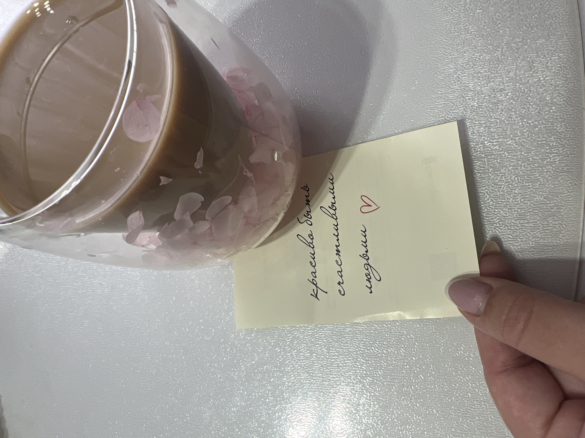 A glass of coffee with flower petals in the liquid is placed on a white surface next to a handwritten note in Russian held by a hand with a polished nail. The note says "красиво быть счастливым любви" followed by a heart. The background is a speckled white, possibly a countertop. The image was likely taken indoors. It is difficult to verify the location as Марковский, Russia from the provided image alone.