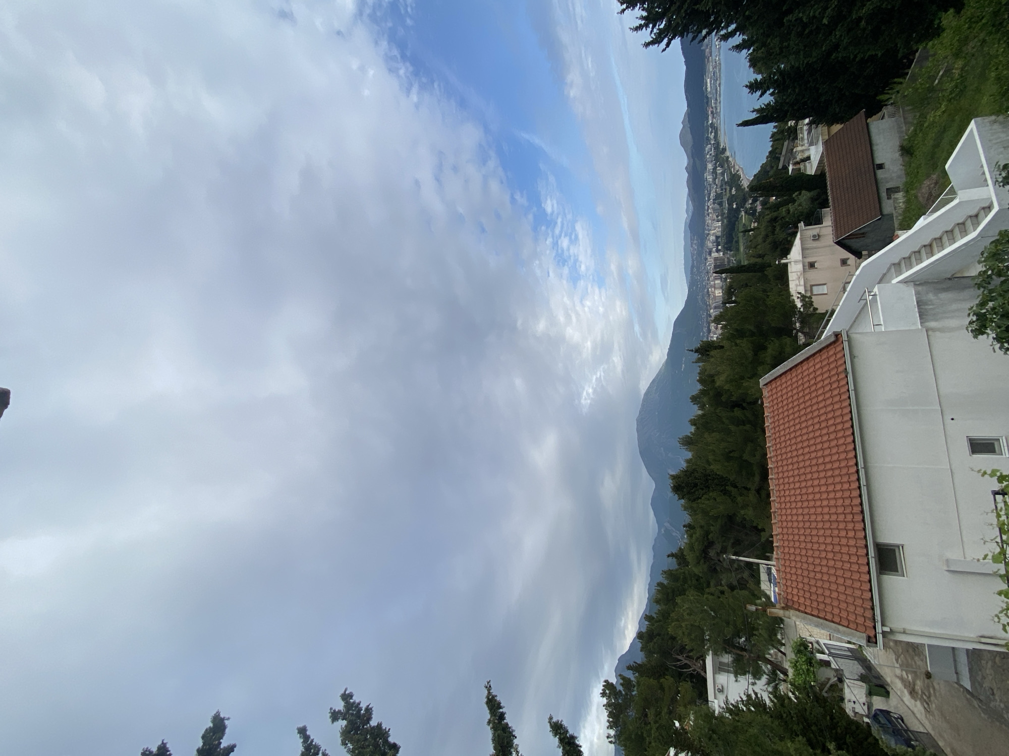 The image shows a view of Šušanj, Montenegro, from an elevated perspective. The sky is partly cloudy with blue patches visible. In the distance, the Adriatic Sea is seen, with the coastline and buildings of the city visible along the shore. A mountain range forms the backdrop. In the foreground, there are trees and several buildings with red-tiled roofs, along with a staircase leading up to a white building.