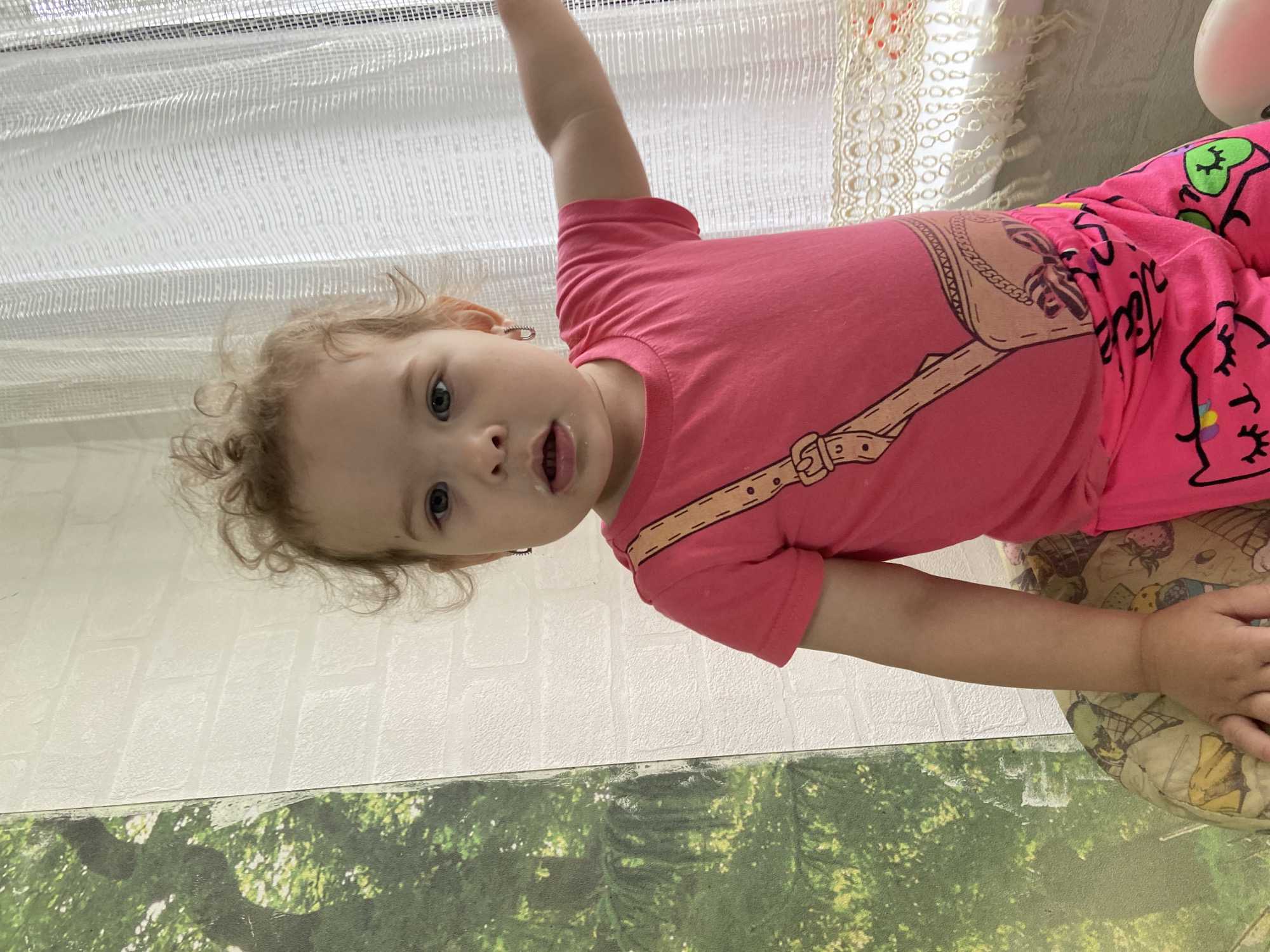 An unknown, young girl with curly blonde hair and blue eyes is depicted from the chest up. She is wearing a pink t-shirt with a purse graphic and pink pants adorned with cat faces. The girl is looking directly at the camera with her mouth slightly open. In the background, a white brick wall and a white curtain with lace trim are visible. To the left of the girl, a large panel with a green forest scene is present. The setting appears to be indoors, possibly a room in a house, with no discernible urban elements to suggest a specific city or location.