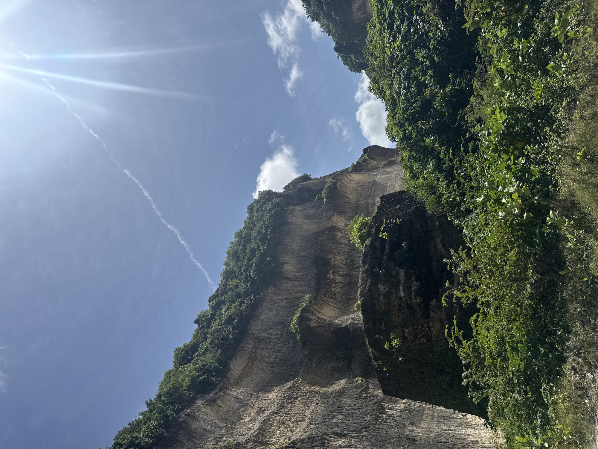 The image shows a cliff face in Kuta Selatan, Indonesia. The cliff is composed of stratified rock, with horizontal layers visible along its surface. Vegetation, including trees and vines, grows along the top and sides of the cliff. The sky is blue with some scattered clouds. A contrail from a plane can be seen in the upper left corner of the image. The sunlight appears to be bright.