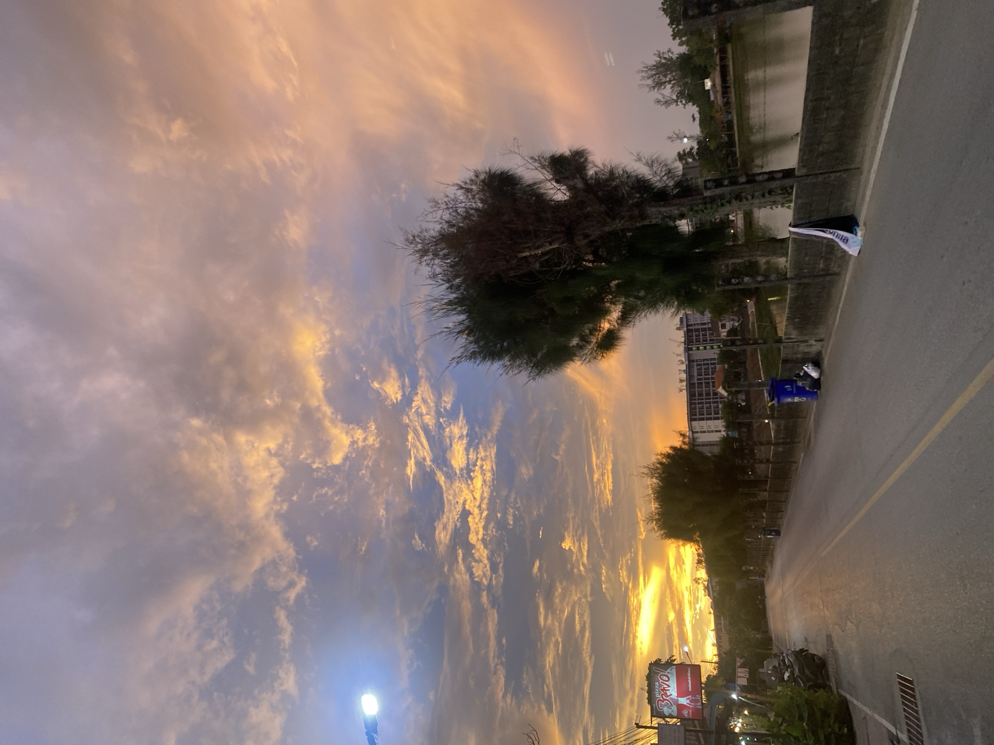 The image depicts a street in Choeng Thale, Thailand, at dusk. The sky is filled with clouds, reflecting orange and gold hues from the setting sun. A road runs along the right side of the frame, marked with a yellow line. A body of water is to the left of the road. The area is lined with trees and buildings. A Pizza Bravo sign can be seen at the bottom of the frame. Streetlights are on, indicating the onset of night.