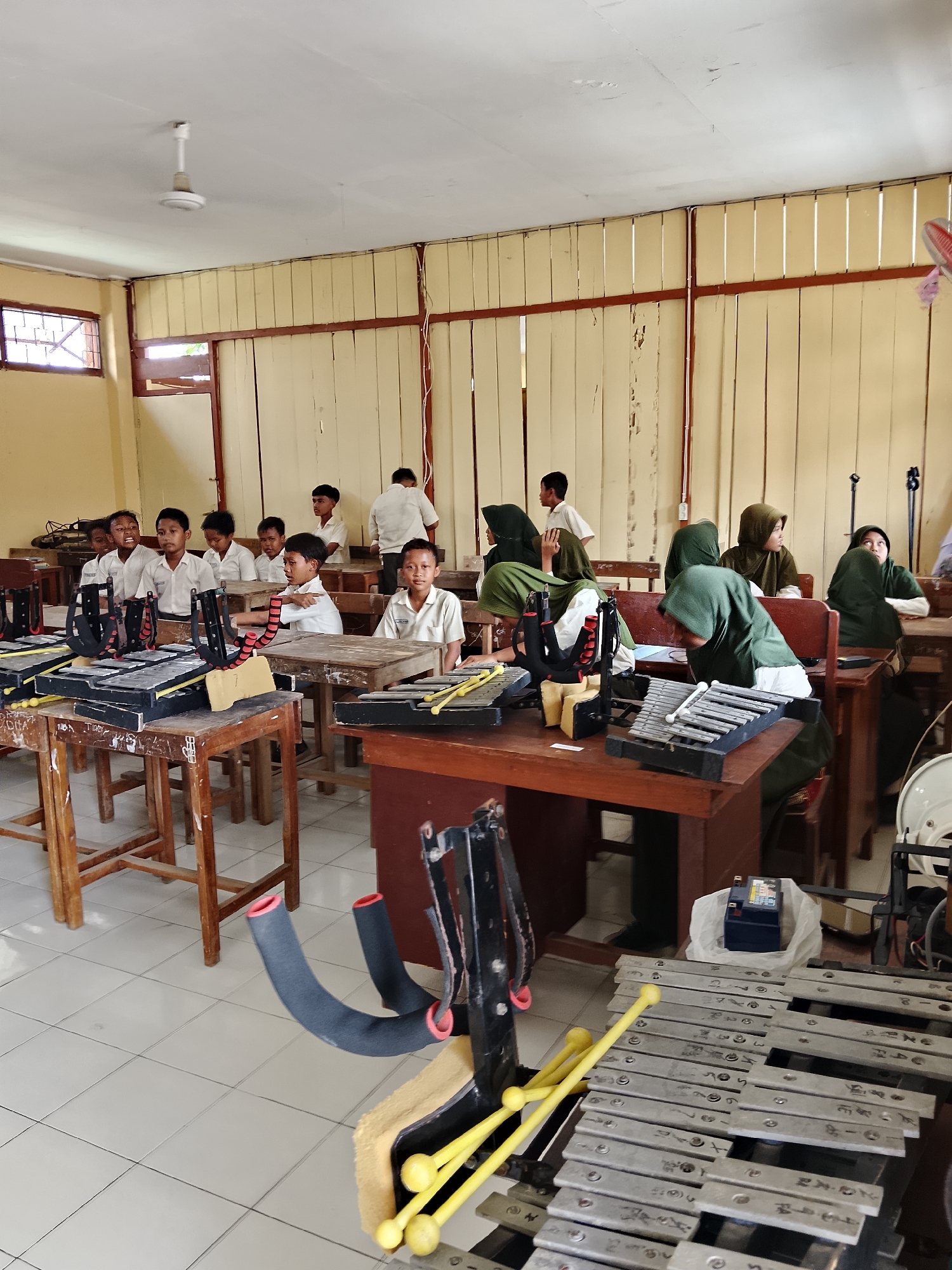 The image shows a classroom in a school, likely located in Kepuh, Indonesia, as indicated by the context. The room is filled with students, many of whom are wearing white uniforms, suggesting they are attending a school in Indonesia. The students are seated at wooden desks, and some of them are looking towards the front of the classroom.
In the foreground and on several desks, there are metallophones, a type of percussion instrument, which suggests that the students might be participating in a music class or a cultural performance preparation. Some of the metallophones have yellow mallets resting on them. The walls of the classroom are painted a light yellow, and there are vertical wooden panels on the upper portion of the walls. A ceiling fan is visible on the white ceiling.
Several students are wearing headscarves, indicating that some of the students are Muslim. The overall atmosphere appears to be one of learning and engagement, with students attentive to an unseen instructor or activity. The classroom is functional rather than modern, with visible signs of wear and tear on the wooden furniture and walls. The lighting suggests it might be daytime, with natural light possibly coming from windows that are not fully visible in the frame.