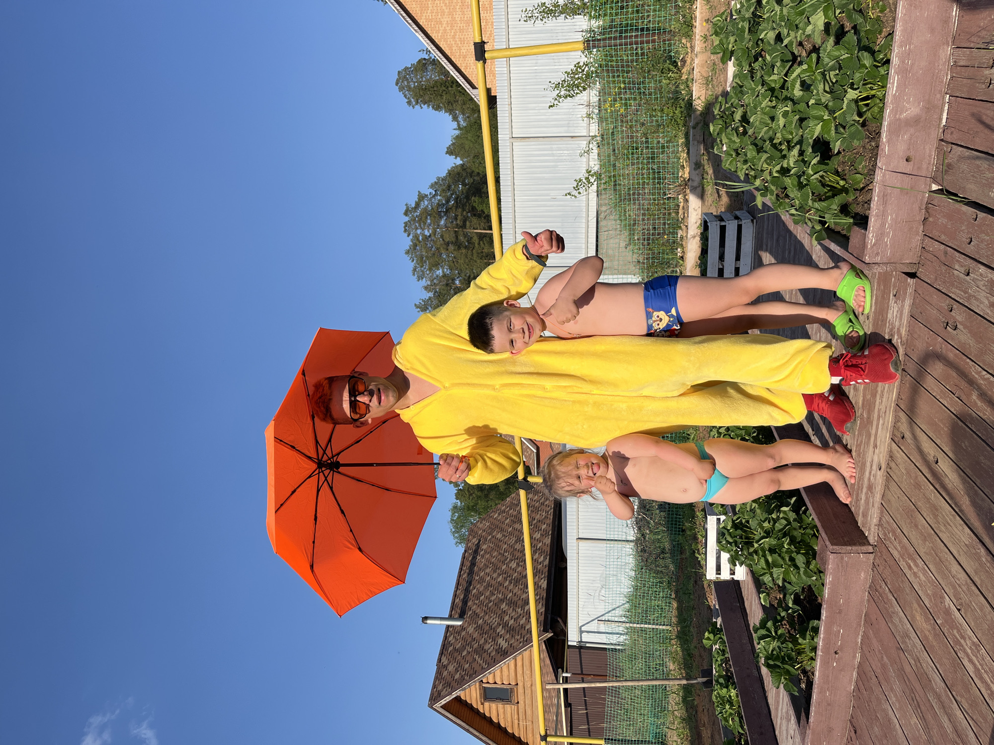 A man wearing sunglasses and a yellow onesie holds an orange umbrella while lying on his back, with two young boys also lying on him. The setting appears to be outdoors, possibly a garden area. The scene includes wooden decking, a wire fence, greenery, and a house in the background. The sky is clear and blue. It is difficult to confirm the location of Chaikovsky, Russia, in the image without further details.