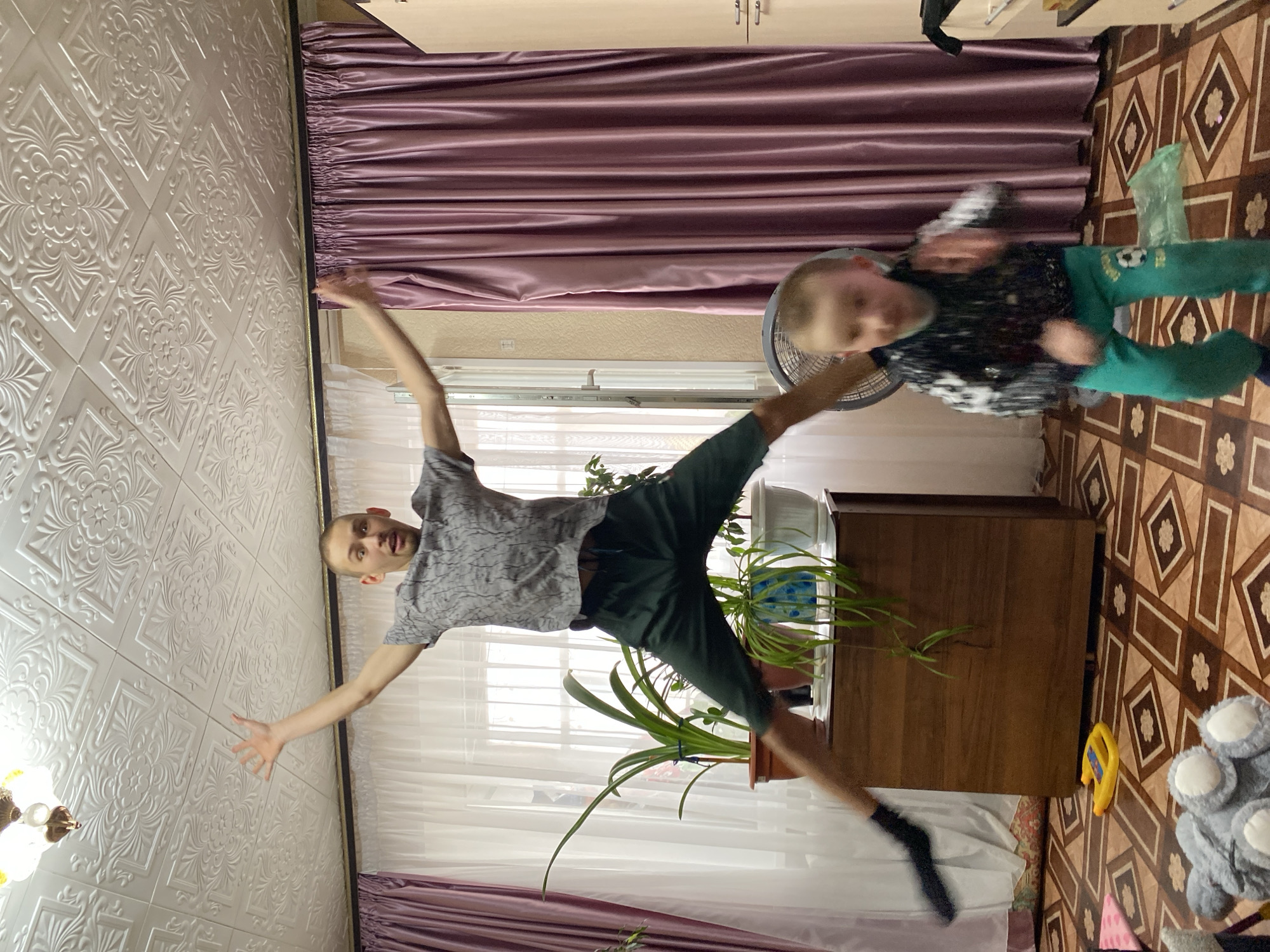 A medium shot, indoor photograph depicts a man and a child in motion within a domestic setting. The man is in mid-air, limbs outstretched as if leaping, positioned in the center. The child, also in motion, appears towards the right side of the frame, seemingly rotating. The room contains a dresser, houseplants, curtains, a fan, toys, and a patterned floor. The photograph's orientation is rotated to the side, as if the camera was held sideways. The location, if in Bely Klyuch, Russia, is inferred from the context of the prompt.