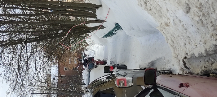 The image displays a winter street scene in Fryazino, Russia. A narrow, snow-cleared pedestrian pathway traverses the center of the frame, bordered on the left by a line of parked vehicles and on the right by a substantial snow bank. The foreground shows a dark gold SUV, identifiable as a Subaru, followed by a red passenger car, both partially covered in snow. The snow bank on the right obscures a dark green metal fence and is topped by accumulated snow. Bare deciduous trees with residual snow cling to their branches are present along the right, one featuring red and white striped warning tape wrapped around its trunk and extending horizontally. In the background, multi-story residential buildings with snow-covered roofs are visible beneath an overcast sky. No human figures are present.