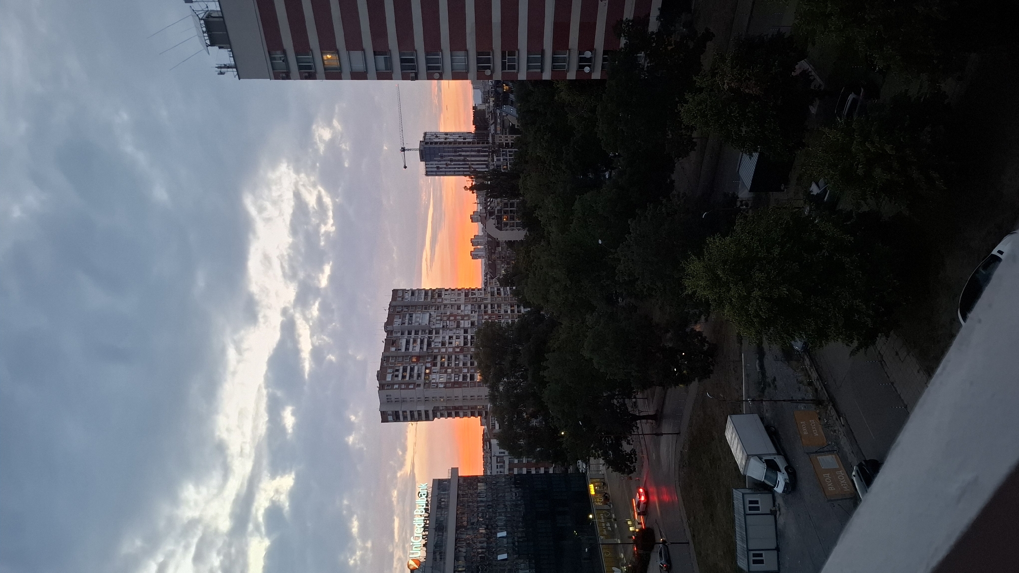 This is an elevated, exterior, daytime, medium-shot photograph taken at dusk, facing east over an urban area in Sofia, Bulgaria, likely from a high-rise apartment. The sky is overcast with scattered clouds, transitioning from a light grey at the top to warmer hues of orange and pink near the horizon, indicating sunset or sunrise.
In the foreground, a white concrete balcony railing occupies the bottom left portion of the image. Further down, a dark grey, slightly wet asphalt road is visible, with parked cars and a white delivery truck. Two portable modular buildings are situated to the left of the truck. Inscriptions in Cyrillic, "ВХОД" and "ИЗХОД" (meaning "Entrance" and "Exit" respectively), are displayed on orange signs. Trees with dense green foliage are scattered throughout the scene, obscuring some of the lower buildings.
Dominating the mid-ground are several multi-story buildings. To the left, a modern glass-fronted building displays the "UniCredit Bulbank" logo. Adjacent to it stands a large, Brutalist-style residential tower block with numerous windows. Further to the right, another tall residential building with red and white horizontal stripes is prominent, featuring a single lit window. In the distance, a construction crane is visible next to a slender, modern high-rise building. The overall impression is of a residential and commercial district experiencing the transition from day to night. There are no people visible in the image.