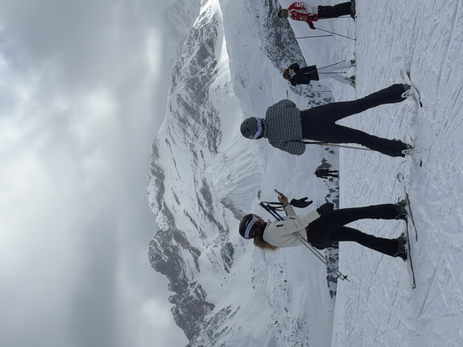The image depicts a snowy mountain landscape on a cloudy day in Courchevel, France. In the foreground, two female skiers are positioned on a groomed ski slope. The skier on the left, with long curly hair, wears a white jacket, black ski pants, and a dark helmet with red and white accents. She holds ski poles in her left hand and a smartphone in her right, appearing to interact with it. Her skis are on the snow. To her right, a second female skier is viewed from the back, wearing a gray patterned top, black ski pants, and a gray helmet with purple accents. She holds ski poles and is also on skis.
In the mid-ground, to the right, three additional skiers are visible. One wears a black jacket and white pants, another wears a red and white jacket (possibly with a Canadian flag emblem) and black pants, and a third, further back, is silhouetted in dark clothing. All are equipped with skis, poles, and helmets.
The background features expansive, snow-covered mountains with visible rock formations under a heavily overcast sky. The overall scene indicates recreational skiing activity in a high-altitude winter environment.