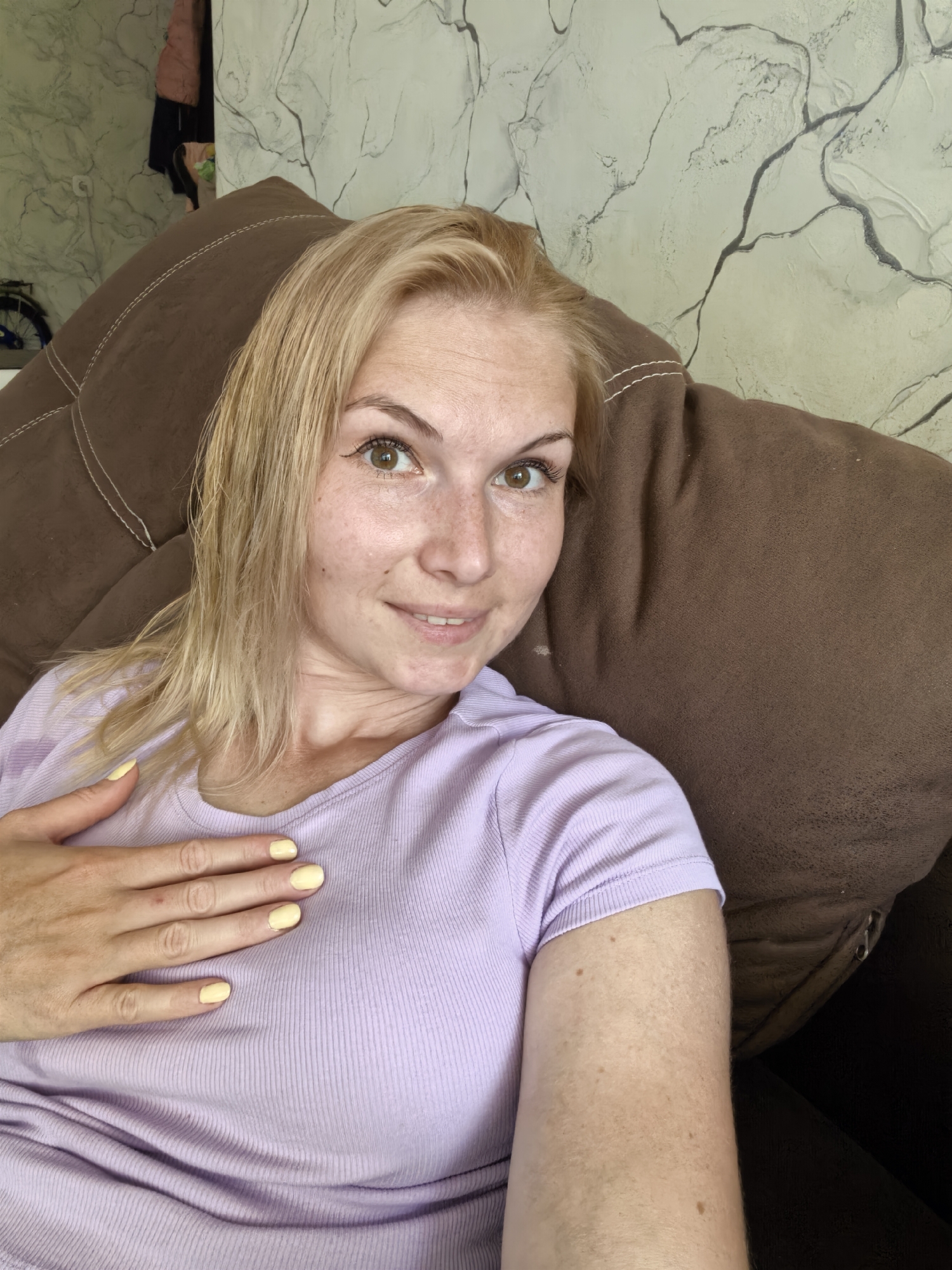 A blonde woman with light skin and freckles is lying on a brown couch, facing the camera with her right arm extended. She is wearing a lavender t-shirt with a ribbed texture. Her left hand is resting on her chest, and her fingernails are painted a pale yellow. Her hair is light blonde and appears wet, falling around her shoulders. The background shows a wall with a textured, marble-like pattern and black line drawings. To the left of the woman, a dark-colored jacket and a bicycle are partially visible. This scene appears to be indoors, likely in a residential setting.