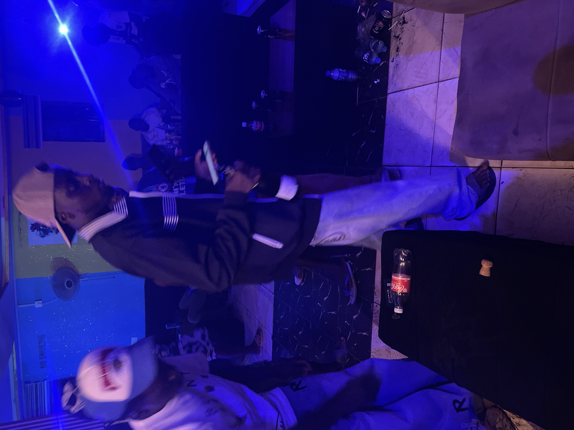 The image depicts an indoor scene in what appears to be a bar or entertainment venue, possibly located in Durumi, Nigeria, given the cultural context implied by the people. The lighting is predominantly blue. Several individuals are present, one holding a mobile phone. Various objects are scattered around including bottles of Coca-Cola. The table is black. A fan is visible on the wall. A "No Smoking" sign is mounted on the wall. The floor is tiled in a cream color with dark accents.
