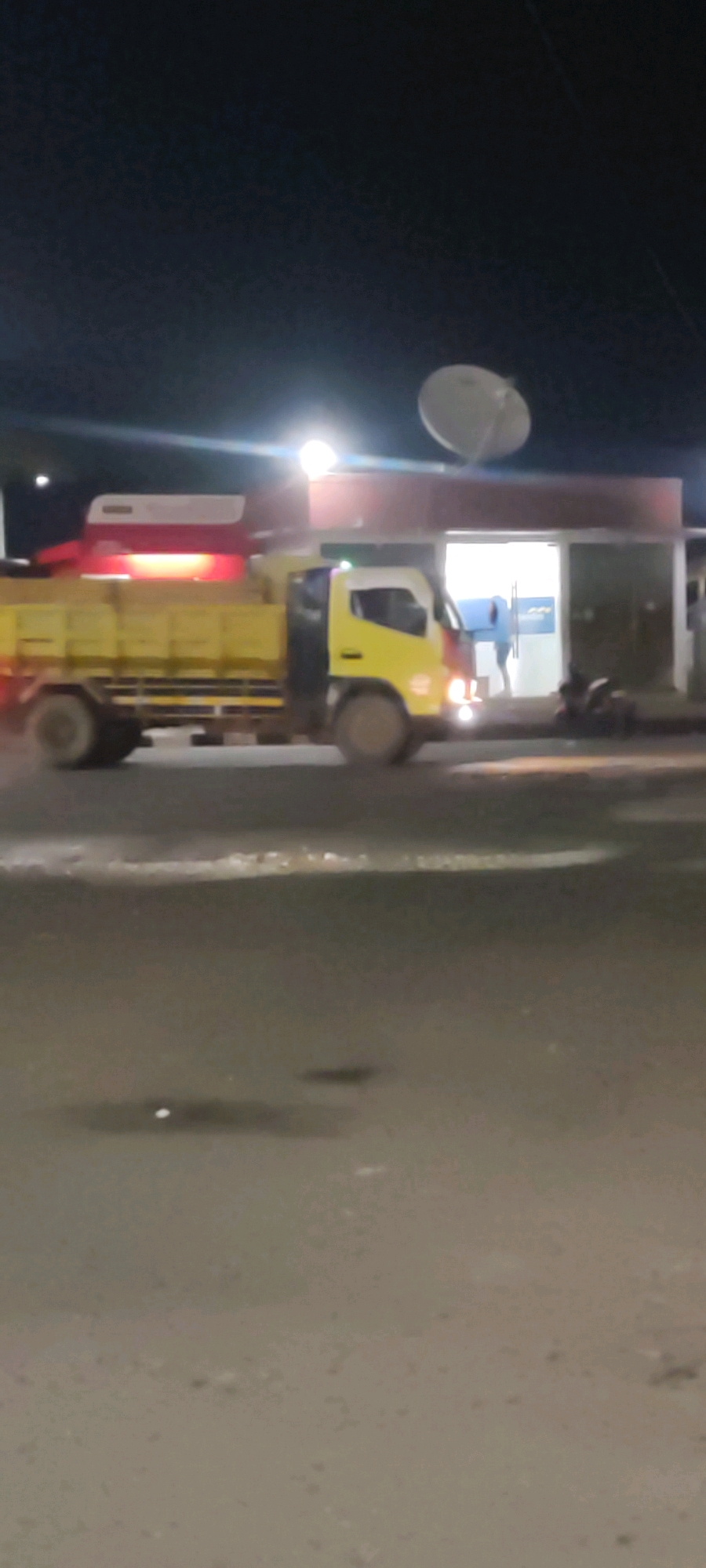 An overexposed, low-resolution, nighttime photograph captures a yellow truck with a red canopy parked in front of a building. The truck's headlights and the building's interior lights are sources of illumination, causing lens flare and blown-out highlights. A large white satellite dish is mounted on top of the building. The building appears to be a commercial establishment, possibly a store or office, with large glass doors. A person in a blue hooded garment is visible inside the building near the entrance. A motorcycle is parked to the right of the building entrance. The foreground shows a dark, wet asphalt road with some reflective patches. The overall scene suggests an urban or suburban environment in Pelayangan, Indonesia, during nighttime.