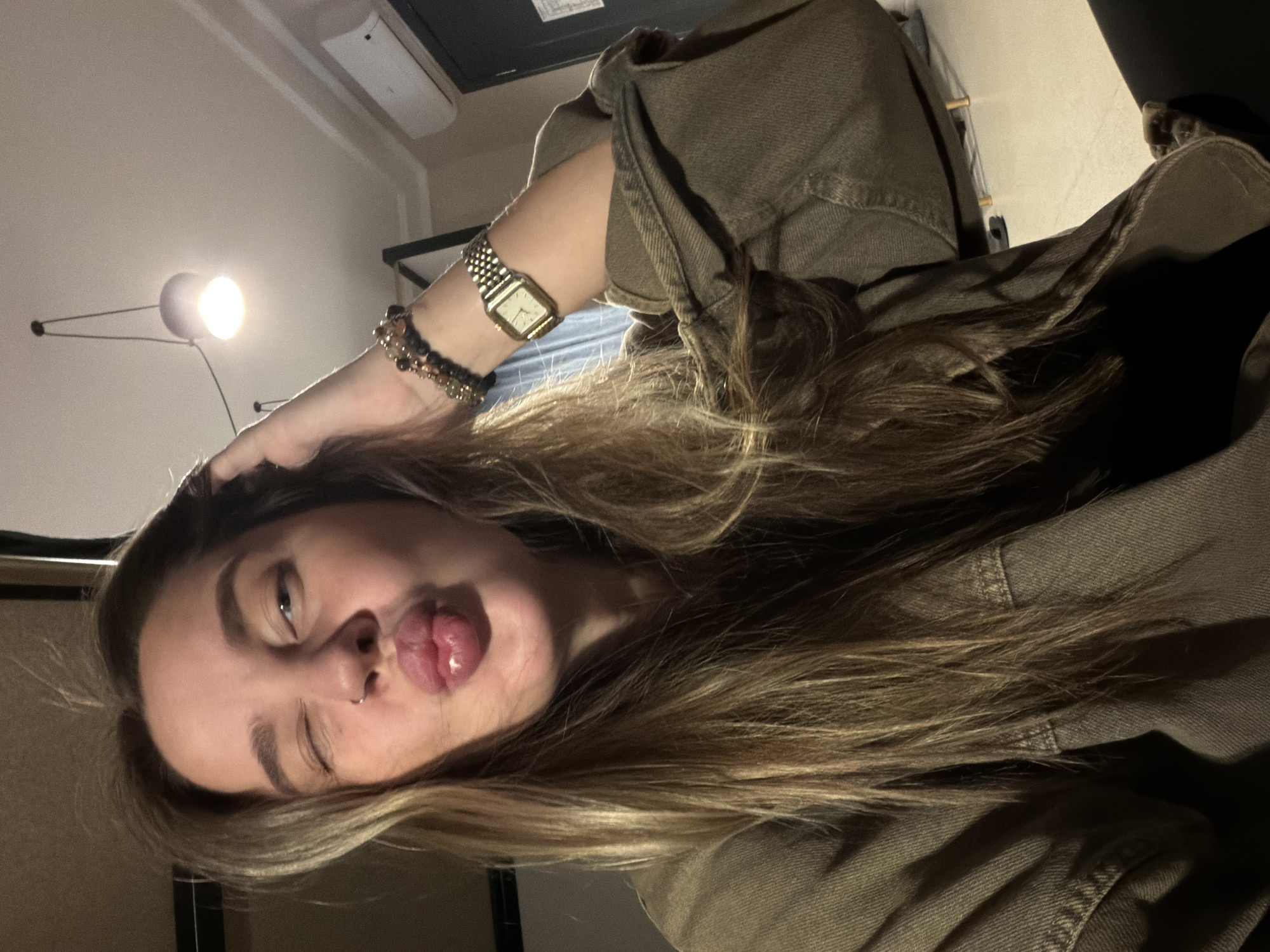 A young woman is captured in a playful selfie, her head tilted dramatically as she winks with one eye and sticks her tongue out slightly. Her long, light brown hair, with visible blonde highlights, cascades over her left shoulder and arm, resting against her olive-green or khaki jacket. She wears a square-faced, gold-colored watch on her left wrist, accompanied by several beaded bracelets in dark and metallic tones. A small septum piercing is visible on her nose.
The setting appears to be an indoor room, likely residential, possibly in Moscow, Russia, given the provided context. A bright, wall-mounted light fixture illuminates the upper left portion of the image, casting a strong glow and creating some shadows. Above it, part of a white air conditioning unit is visible on the ceiling. The background is otherwise minimally detailed, suggesting a simple wall and possibly dark furniture or shelving. The overall lighting is artificial and relatively dim outside the immediate bright area. The image is oriented diagonally, adding to its dynamic and informal feel. No discernible text is visible within the frame.