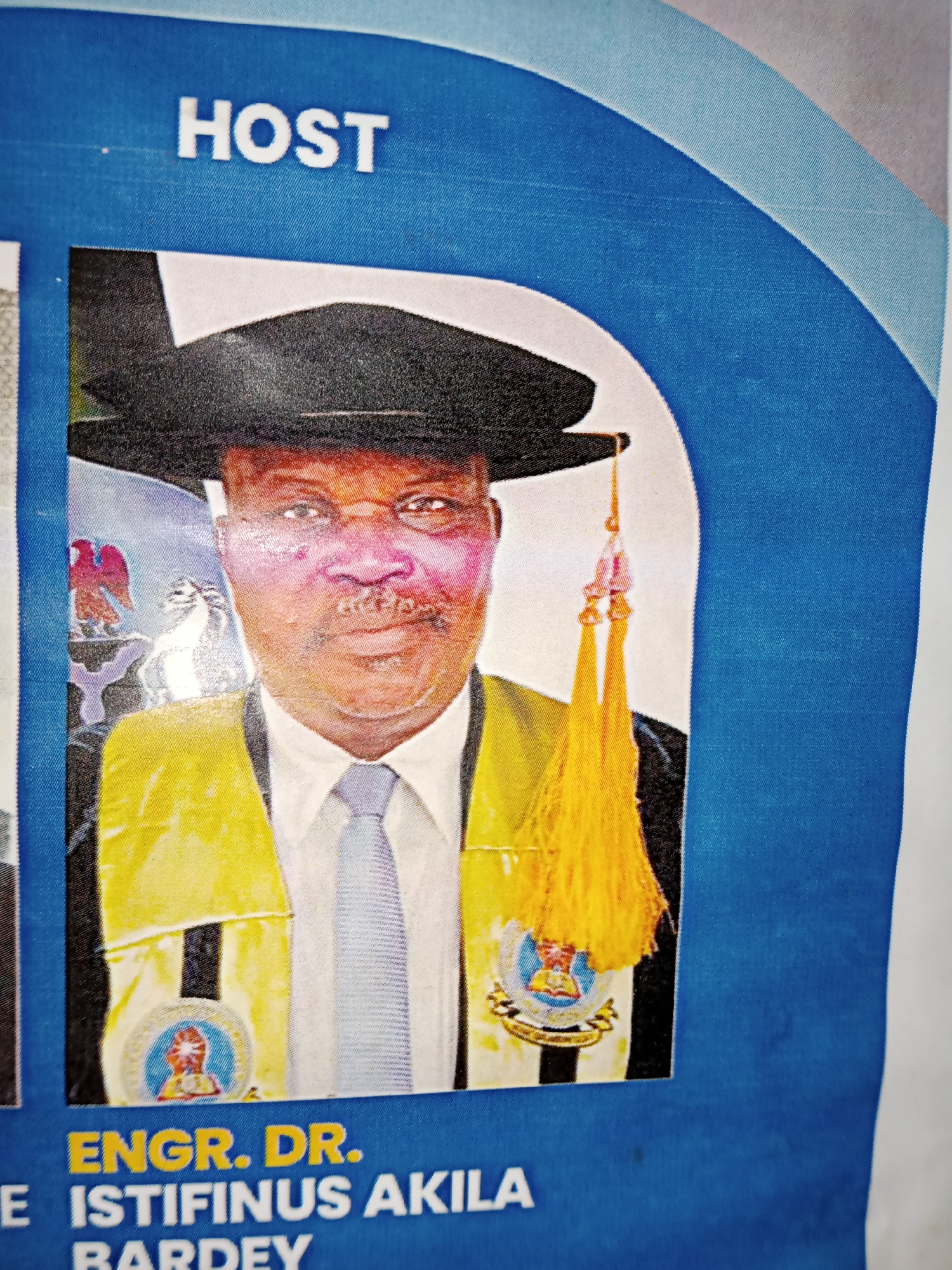 The image shows a portion of a large poster or banner, likely displayed in Jalingo, Nigeria, given the provided context. The dominant colors are blue and white, with vibrant yellow and other colors in the central portrait.
At the top of the poster, the word "HOST" is prominently displayed in white, block capital letters against a blue background. Below this, a portrait of a man wearing academic regalia fills the central space. He is adorned with a black academic cap, a yellow sash over a white shirt, and a patterned tie. A decorative yellow tassel hangs from his cap. Behind him, the image contains elements that appear to be from official seals or emblems, including a bird of prey and what looks like a coat of arms.
Below the portrait, text in bold, yellow capital letters reads "ENGR. DR." Below this, in white capital letters, is the name "ISTIFINUS AKILA" and then "BARDEY". The background of this text area is a deep blue, with a curved, lighter blue border framing the portrait area.
The overall impression is that of an official announcement or program for an event, likely a graduation ceremony or an academic conference, where the individual named is a host or a distinguished guest. The image is of moderate quality, with some visible texture from the material of the banner and minor imperfections like smudges.