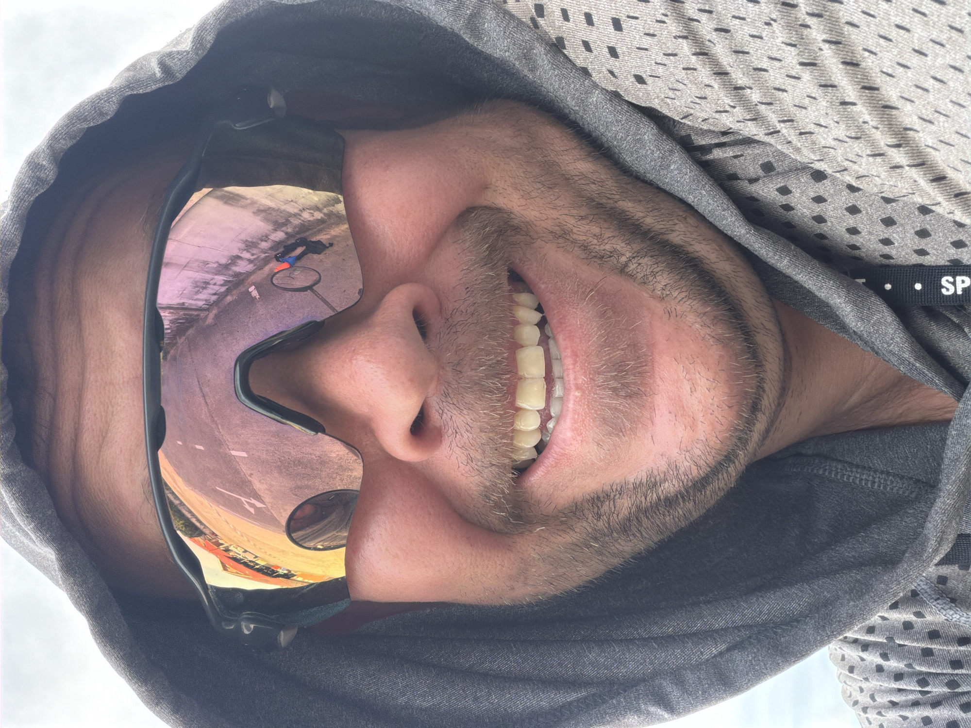 A man wearing a gray hooded jacket and sunglasses is smiling. The sunglasses reflect a street scene with buildings and a bicycle. The location is identified as Baan Mae Nam, Thailand.