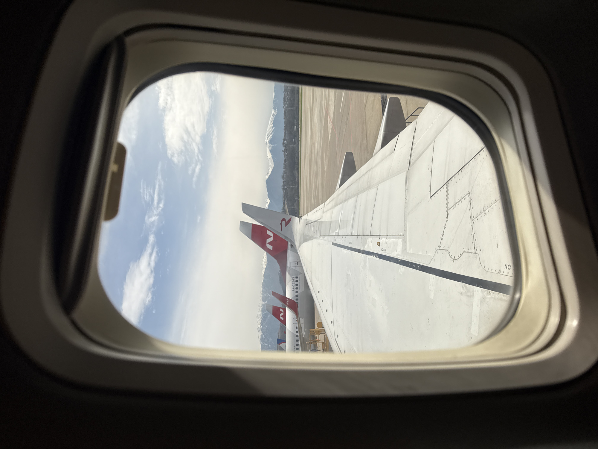The image shows a view from an airplane window. Visible outside is the wing of the plane and other airplanes parked nearby. In the background are mountains under a partly cloudy sky. The tail fins of the adjacent planes have the letter "N" on them. The airplane appears to be on the ground. Without additional information, it's impossible to confirm the specific location is Sochi, Russia.