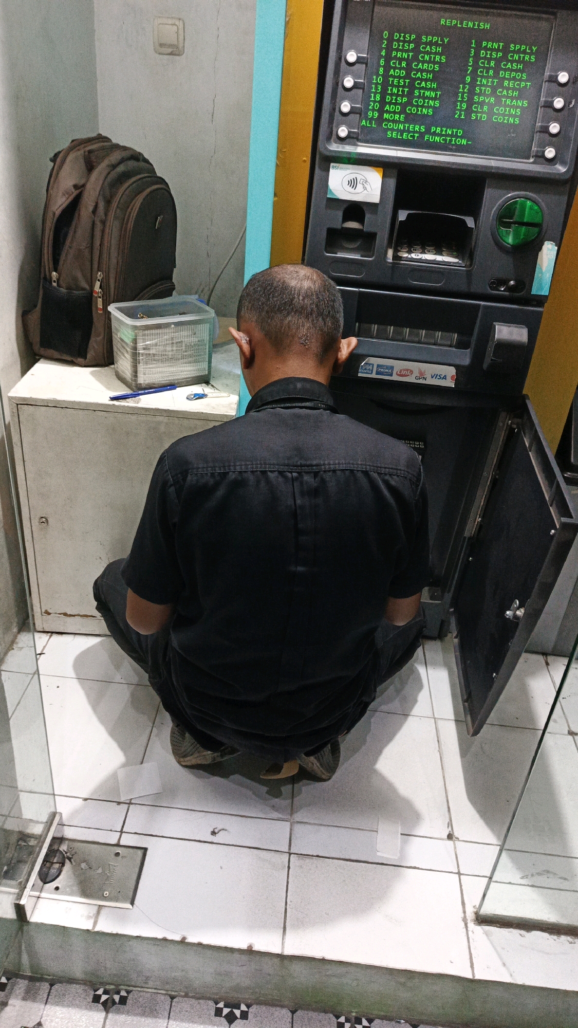 A man in a black uniform is crouched in front of an ATM in Gondangrejo, Indonesia. The ATM screen displays a list of functions, including "DISP SPLY," "DISP CASH," and "ADD CASH." The man appears to be interacting with the ATM, possibly performing maintenance or a transaction. To the left of the ATM, a brown backpack is placed on a white cabinet. The environment appears to be an indoor public space, possibly a bank or a retail store, with tiled flooring and a plain wall. The lighting is dim, suggesting it might be evening or an indoor setting. The overall scene depicts a routine or perhaps an unusual interaction with an ATM.