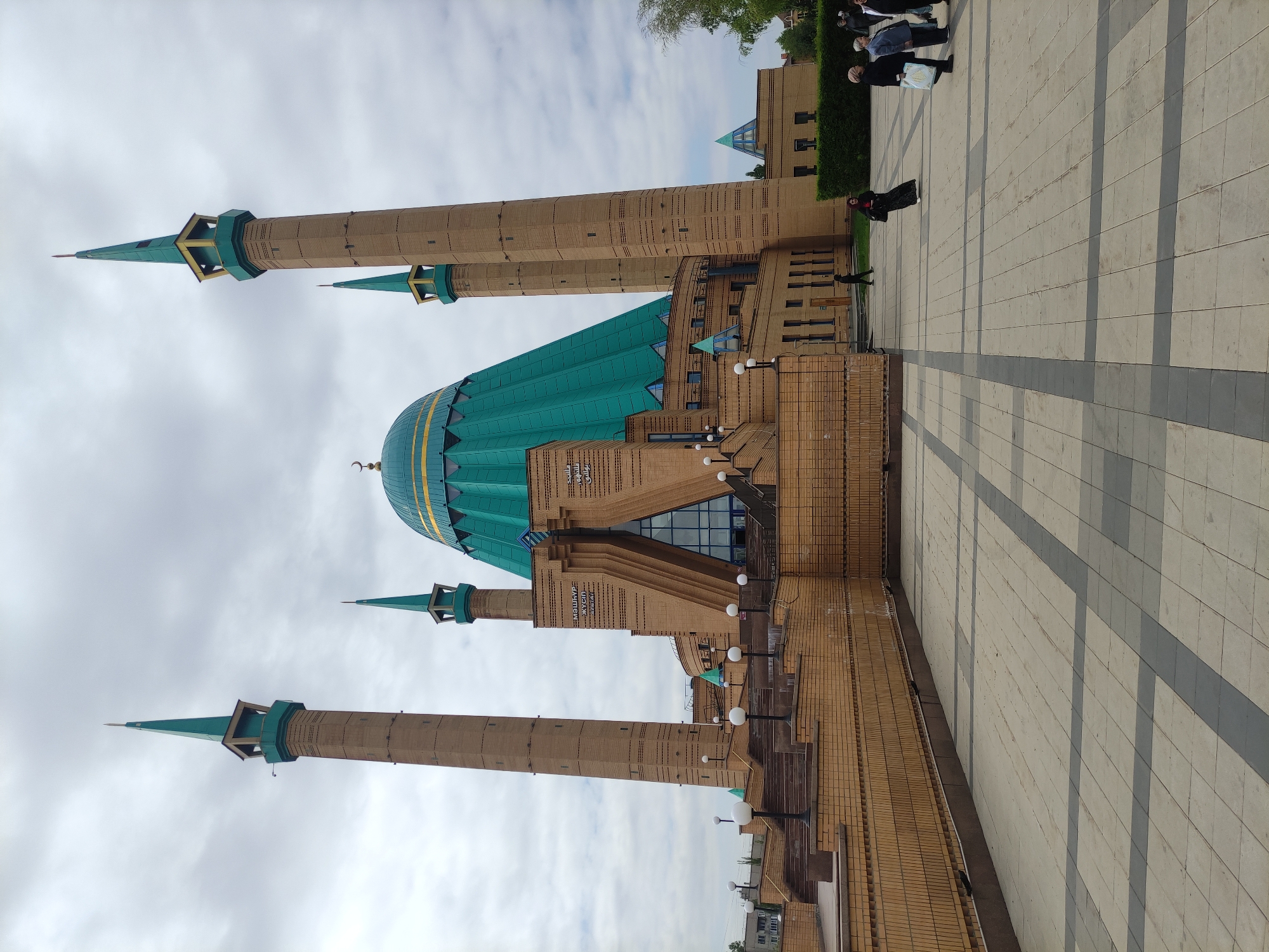 The image shows the Kul Sharif Mosque in Kazan, Russia, not Pavlodar, Kazakhstan. The mosque features a large turquoise dome and four minarets made of tan brick. The sky is overcast with light gray clouds. Several people are seen walking on the paved plaza in front of the mosque.
