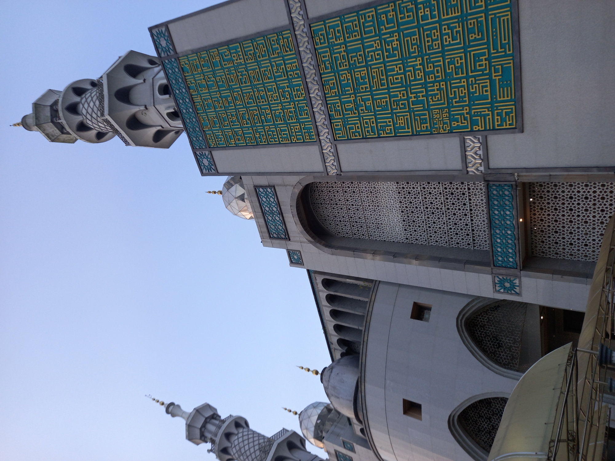 A low-angle, full-frame, eye-level outdoor shot captures the exterior of the Masjid Raya Samarinda, located in Samarinda, Indonesia. The sky is a clear, pale blue. The mosque's architecture is characterized by a tall, multi-tiered minaret on the right, adorned with geometric patterns and Islamic calligraphy in green and gold. To the left, another, smaller minaret with a golden finial is visible. The mosque features domes, arched windows with intricate latticework, and a gray, stone-like facade. In the foreground, a portion of a covered walkway or canopy with a metal frame is visible. No people are present in the image.