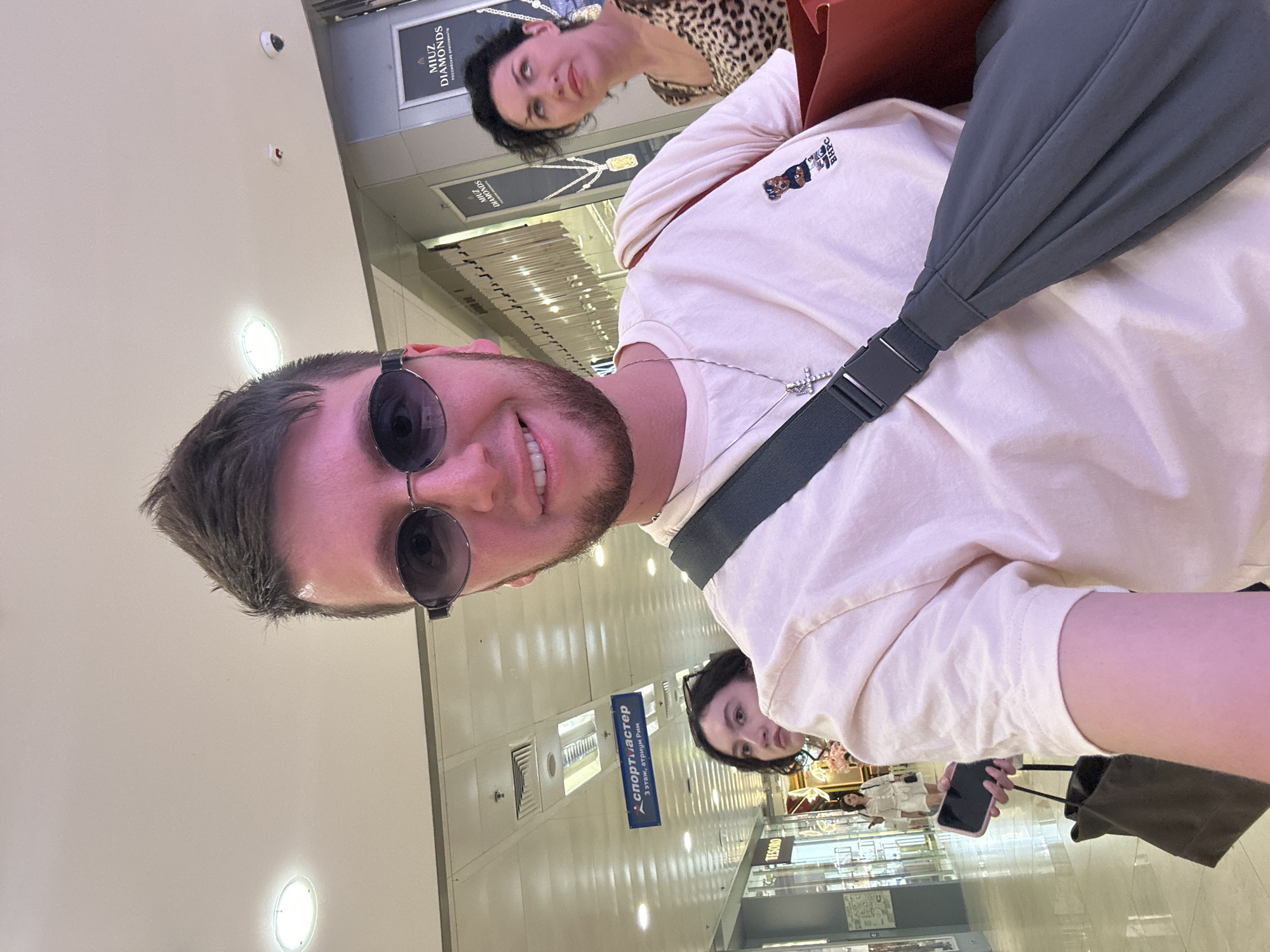 In Moscow, Russia, a young man with spiky brown hair and sunglasses takes a selfie indoors, likely in a shopping mall. He wears a light-colored t-shirt with a small graphic of a bear and the letters "BHPC" on the left breast, and a silver cross necklace. A dark gray sling bag is worn across his chest. In the background, a woman with dark curly hair wearing a leopard-print top looks up and to the right, and another woman with dark hair is partially visible in the mid-ground, looking to the left while holding a phone. Various store signs are visible, including "СПОРТМАСТЕР" (Sportmaster), "TESORO," and "MIUZ DIAMONDS." The environment appears to be a brightly lit, indoor walkway with a tiled floor and ceiling lights.