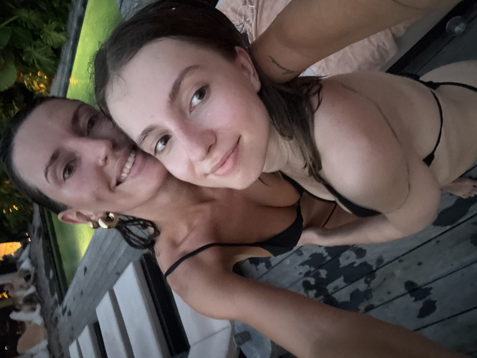 Two women are depicted in a close-up, outdoor portrait taken at an evening or nighttime poolside setting in Canggu, Indonesia. Both subjects have wet, dark hair and are wearing black string bikinis. The woman on the left, who appears slightly older, has her hair pulled back and is smiling directly at the camera. She wears large gold-colored hoop earrings and a nose piercing. Her right arm is partially visible, likely holding the camera or extending towards it. The woman on the right, appearing younger, has her hair pulled back and is smiling subtly, looking slightly towards her right. Her left arm is wrapped around the waist/hip area of the woman on the left. A tattoo reading "luna" is faintly visible on her left inner forearm. The immediate foreground shows a wet wooden deck. In the background, a swimming pool with illuminated, greenish water is visible, bordered by lush, illuminated tropical foliage. Several light-colored pool loungers are partially visible on the deck to the left of the subjects. A light-colored fabric is visible on the far right.