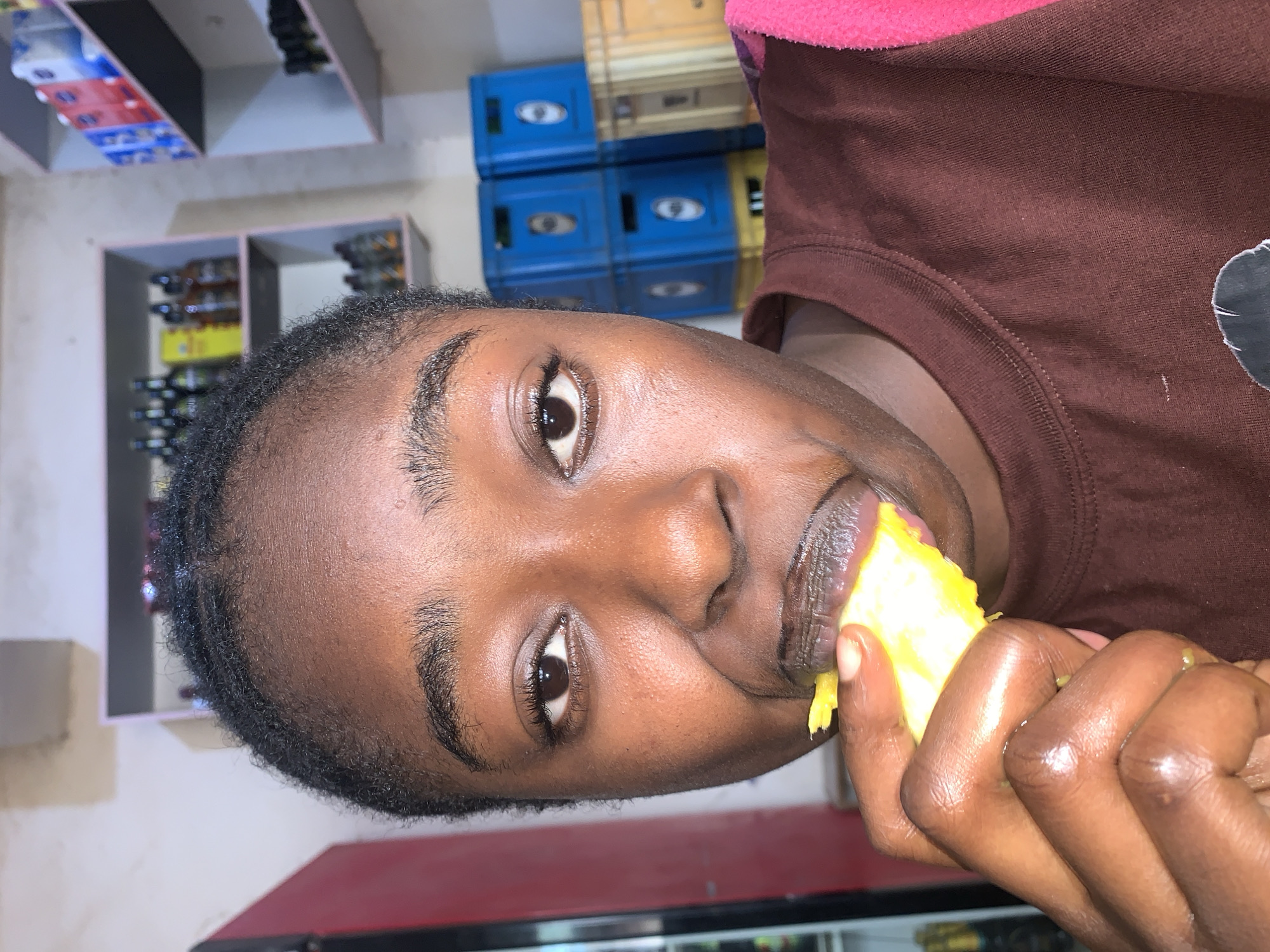 A close-up shot shows a person with brown skin eating a bright yellow piece of food. The individual is wearing a brown long-sleeved shirt with a pink collar. The person is looking at the camera. In the background, there are shelves stocked with bottles and containers, and blue plastic crates stacked on top of each other. The photograph was taken in New Stadium, Nigeria.