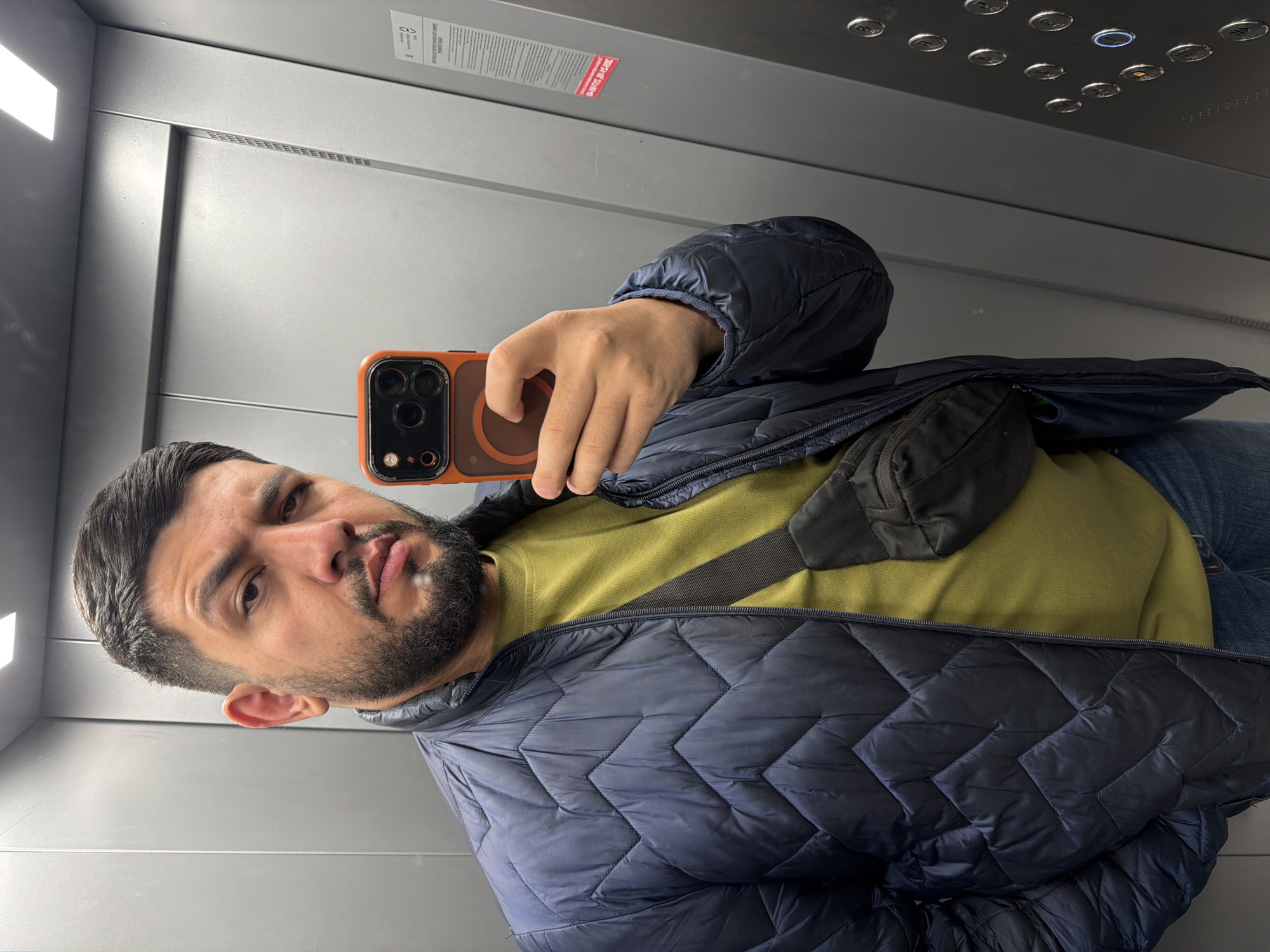 A man is taking a selfie in an elevator. He is wearing a yellow shirt and a dark blue quilted jacket, with a black bag slung across his chest. He holds an orange cell phone in his right hand. The elevator has a gray interior, with buttons visible at the top and a notice fixed to the wall. The location is likely in Krasnoyarsk, Russia, as per the prompt's request.