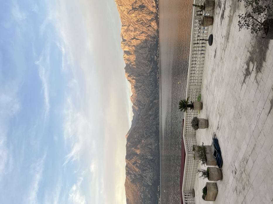 The image depicts a waterfront scene in Donji Orahovac, Montenegro, under a partly cloudy sky with ambient lighting suggesting either sunrise or sunset. The foreground features a tiled outdoor terrace constructed from light-colored square paving stones. Several potted plants of varying sizes and species are arranged across the terrace; these include cylindrical and rectangular planters containing small shrubs and a larger palm-like plant. A dark blue inflatable boat or similar watercraft lies flat on the tiles. A white balustrade with ornate spindles runs horizontally, separating the terrace from a large body of water, likely the Bay of Kotor. Behind the balustrade, a red cushion or mattress is visible on a lower ledge. On the water, a single yellow buoy and several distant boats are discernible. The mid-ground is dominated by the calm, greyish water, which reflects parts of the sky and the distant mountains. The background is composed of a massive range of rugged, rocky mountains, with their peaks and upper slopes illuminated by sunlight, exhibiting shades of ochre and grey, contrasting with shadowed ravines. The sky above is light blue with scattered, thin cirrus clouds. No individuals are present in the scene.