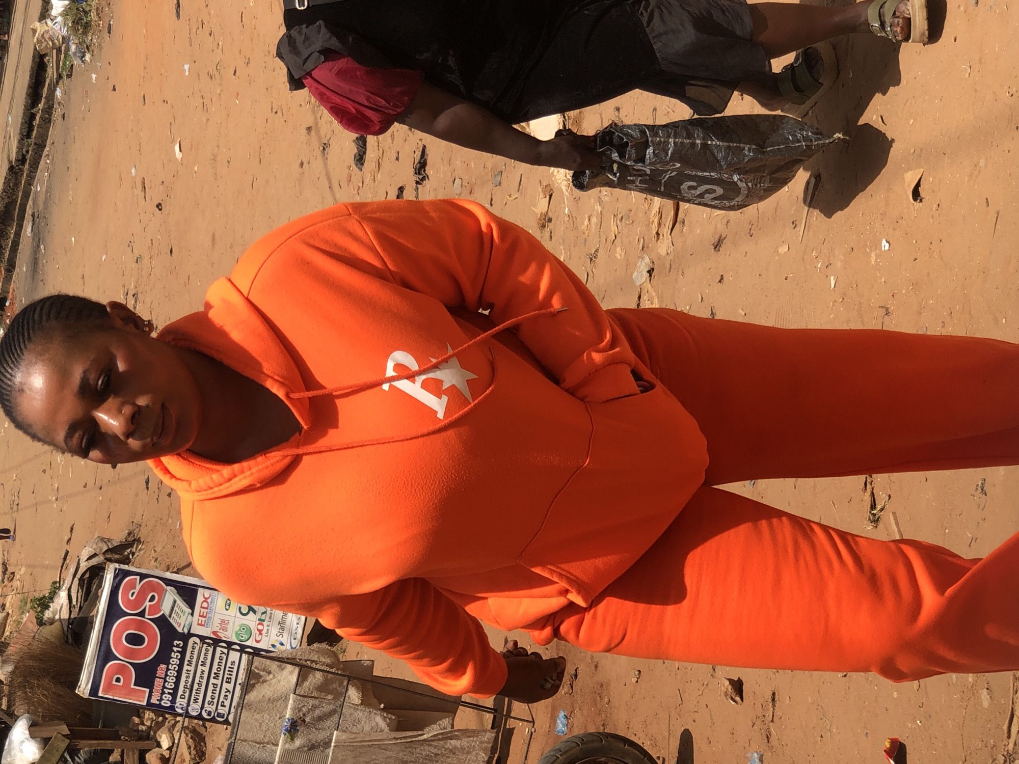 The image shows a woman in an orange tracksuit standing on a sandy street in Onitsha, Nigeria. She is facing the camera, with a POS (Point of Sale) sign nearby that lists services such as depositing and withdrawing money, sending money, and paying bills. The sign also advertises local telecommunication and utility companies. Another person is partially visible in the background, wearing dark clothing and holding a plastic bag. The ground is sandy and strewn with debris.