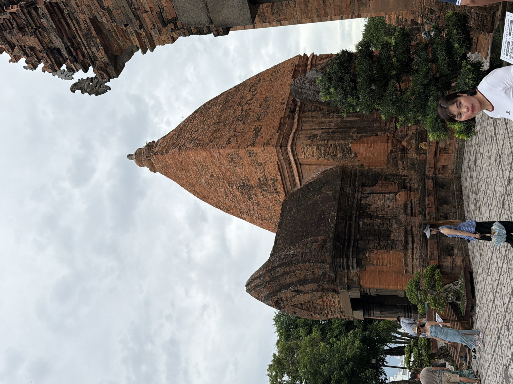 This image shows the Po Nagar Cham Towers in Nha Trang, Vietnam, a historical site constructed from red brick. The sky is overcast with clouds. In the foreground, a woman wearing a white t-shirt with a red trim is looking towards the camera. To her right is a large, well-maintained evergreen tree. Behind her, several other people are visible. One woman in a blue and white checkered top and a grey sun visor stands holding a closed umbrella. Another woman in a light blue dress with a sun hat is walking on the paved ground near a set of stairs. A person in a grey shirt is visible on the stairs. Two men are standing further in the background to the left, one wearing a grey t-shirt and shorts. Bonsai trees are strategically placed around the paved courtyard. The architecture of the towers is characterized by their pyramid-like structures and intricate brickwork.