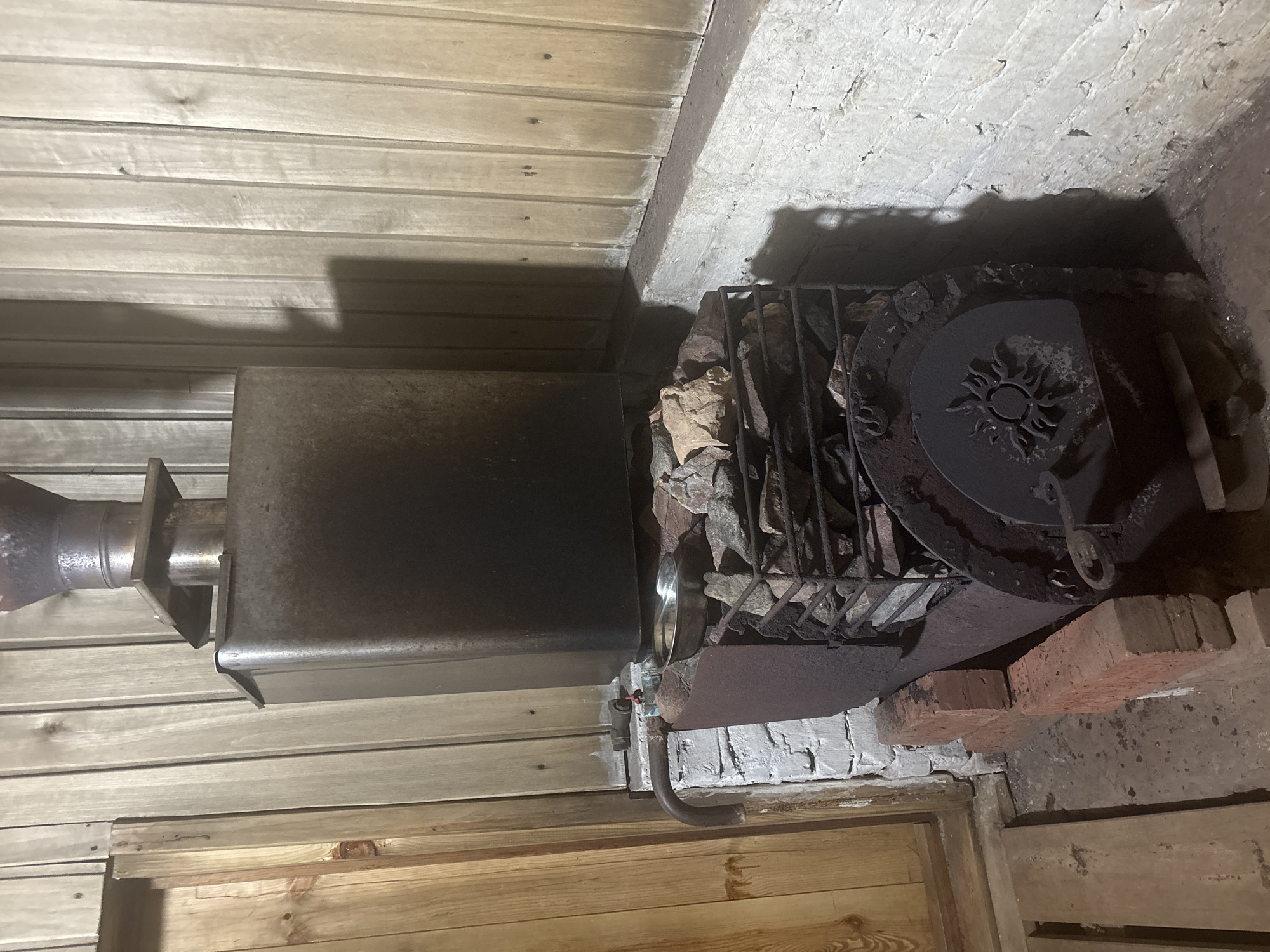 A wood-burning stove is positioned in the corner of a room with wooden walls. The stove is constructed of metal, and is covered with rocks. A metal pipe, potentially a flue, extends from the stove. The scene is located in Chaikovsky, Russia.