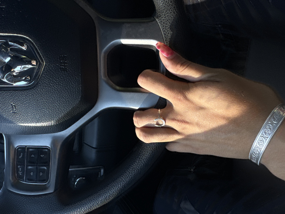 The image depicts a close-up, first-person view from inside a vehicle, focusing on the steering wheel and a human left hand. The steering wheel is black with silver-colored spokes, featuring a prominent chrome Ram truck logo in the center and "SRS AIRBAG" embossed below it. Control buttons for audio and phone functions are visible on the left spoke. A medium-toned human left hand rests on the lower right spoke of the steering wheel. The hand wears a silver-colored horseshoe-shaped ring on the ring finger and a wide, silver-colored bangle with an ornate patterned design on the wrist. The visible thumb has a red/pink, possibly marbled, fingernail. Strong sunlight illuminates the hand and a portion of the steering wheel, casting shadows. The specific location within Rancho Cordova, United States, is not discernible from the visual information provided.
