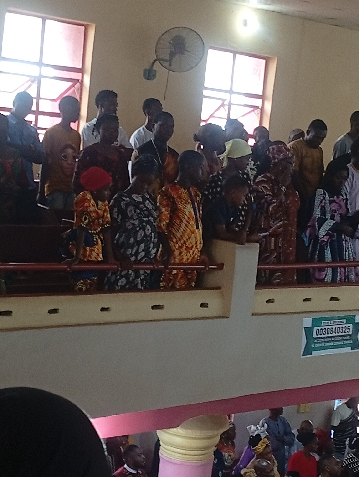 A church service is underway, with a crowd gathered on an upper level within a building. Two windows let natural light into the room. A mechanical fan hangs from the ceiling. A sign displaying the text "0030840325" is fixed to the balcony railing. Various people wearing different clothing are visible, some looking down at the scene below.