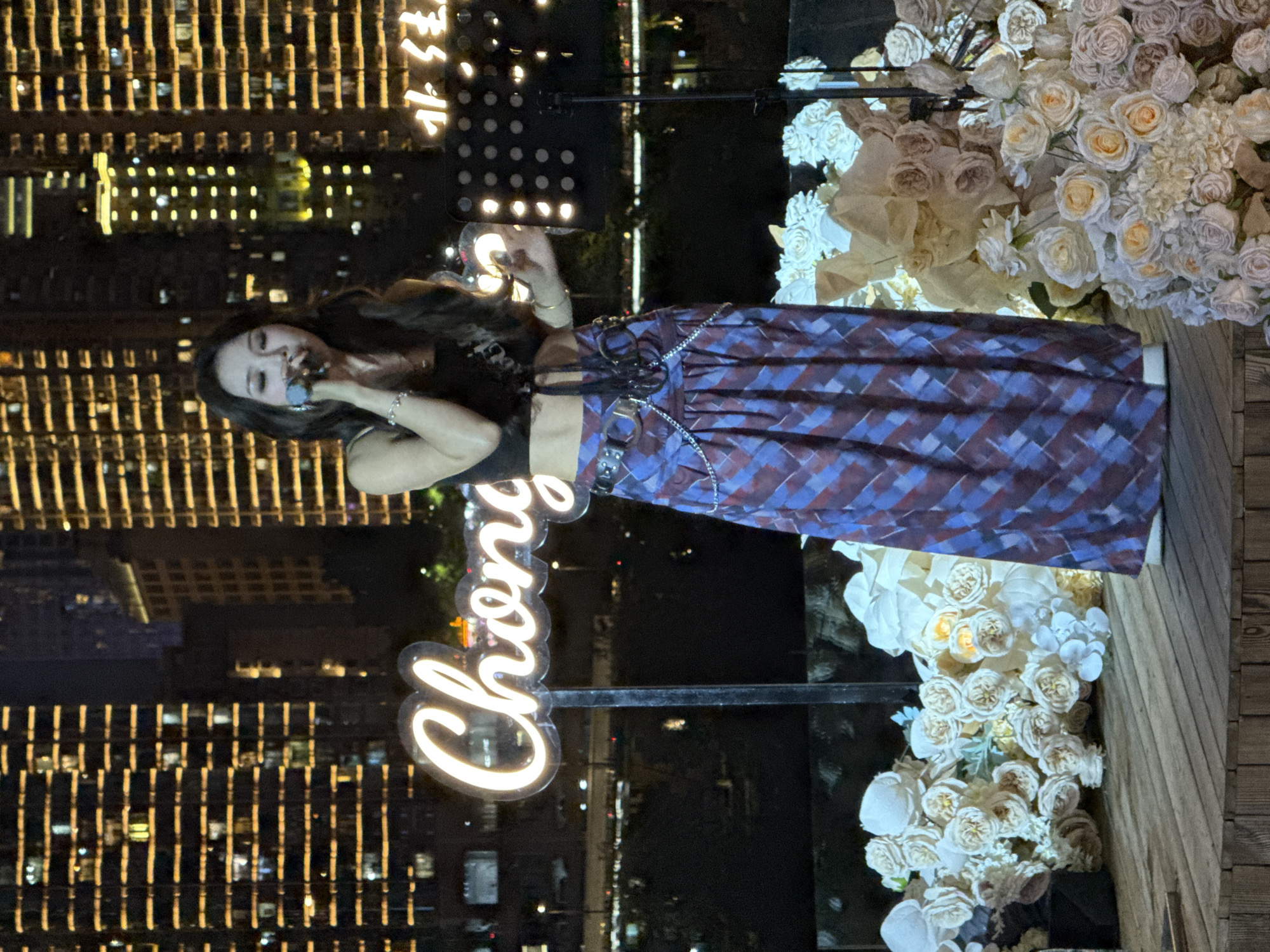In the foreground of the image, a woman stands on a wooden stage, holding a microphone to her lips as if singing. She wears a dark, sleeveless top and a long, plaid skirt in shades of blue and red, cinched at the waist with a wide belt featuring a chain accessory. She is adorned with bracelets and earrings. Surrounding the stage are numerous arrangements of artificial white and cream-colored flowers, some with hints of yellow and blue.
Behind the stage, a neon sign with the word "Chong" in a cursive font is prominently displayed. Further in the background, towering buildings with countless illuminated windows create a cityscape, suggesting an urban environment. The overall scene is set at night, with the city lights reflecting and contributing to the ambient lighting. Based on the neon sign and the architectural style of the buildings, the location is identified as 江北城街道, China.