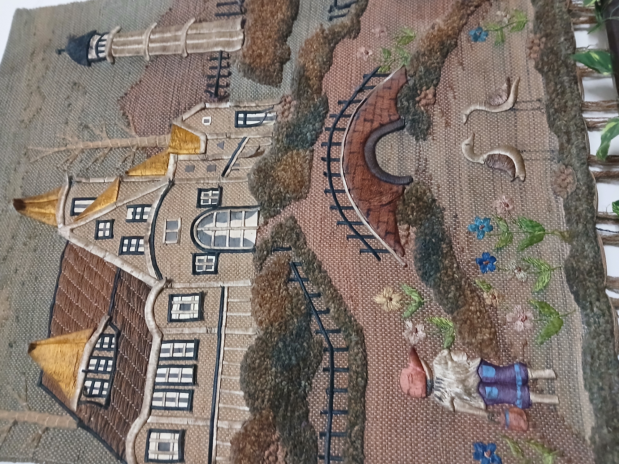 This media file showcases a detailed textile art piece, possibly an embroidered or appliqué tapestry, depicting a whimsical village or town scene. The image was taken in Bengaluru, India.
**Subjects:** The artwork features a stylized human figure, resembling a child, lying on its back in the lower right portion of the scene. The figure is dressed in blue shorts and a light-colored top, with a reddish hat or hair. Two light-colored, slug-like or worm-like creatures are also depicted on the ground nearby.
**Objects:** The scene is rich with various embroidered objects. Prominently, there are several multi-story buildings, likely houses, with intricate window details, dark outlines, and varied roof colors (brown and golden-yellow). One tall, cylindrical structure resembling a lighthouse with a dark cap stands in the upper left. A reddish-brown arched bridge with black railway tracks spans a gap in the landscape. Numerous trees and shrubs, rendered with textured stitching, are scattered throughout, giving a sense of foliage. Small, delicate embroidered flowers in shades of blue, pink, and yellow dot the ground.
**Scene and Setting:** The textile art portrays an idyllic outdoor landscape. The background fabric has a coarse, woven texture, possibly burlap or a similar material. The setting appears to be a quaint village or rural area, characterized by its distinctive architecture, including gabled roofs and varied window designs. The presence of a railway and bridge suggests connectivity within this whimsical world.
**Activity or event taking place:** As a static art piece, there is no active event taking place. The scene captures a peaceful moment, with the human figure resting amidst the natural and architectural elements.
**Notable details:** The artwork exhibits a high level of craftsmanship with different stitching techniques, creating texture and depth. The gold-colored roofs of some buildings catch the light, adding a subtle shimmer. The weather appears fair and bright, implied by the outdoor scene and colors, suggesting daytime. The architectural style has a generic, storybook feel, not strongly tied to a specific real-world geographical location within the artwork itself, although the image was captured in Bengaluru, India. The overall impression is one of charm and tranquility. There is no visible text within the artwork.