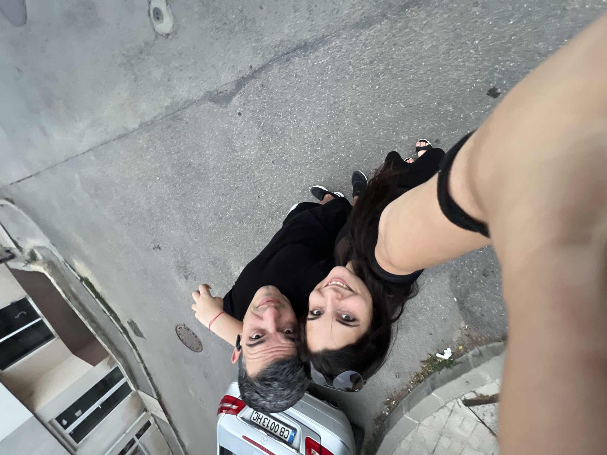A man and a woman are taking a selfie outdoors. They are both wearing black clothing. The man has graying hair and the woman has long dark hair. They are standing on a paved street, and a silver car with a Bulgarian license plate is parked nearby. The background shows a building with windows and a sidewalk. The image appears to have been taken during the day. Based on the license plate format "CB 0013 HC", this indicates a Bulgarian license plate, likely from the city of Varna, not Sofia, as these plates are registered in the Varna province.