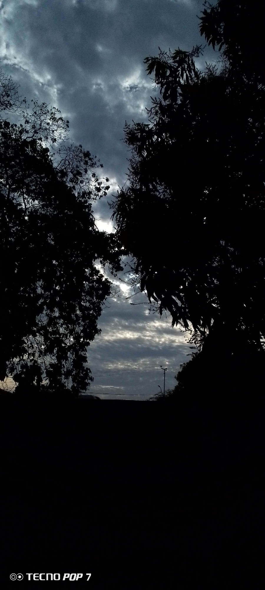 The image shows a sky filled with dark, layered clouds in Altagracia, Venezuela. The view is framed by silhouetted trees on either side, with the trees appearing darker due to the low lighting conditions. A distant utility pole and some buildings are visible through the gap in the trees beneath the cloudy sky. The foreground is predominantly dark and indistinct. The brand name "Tecno Pop 7" is present at the bottom of the image.