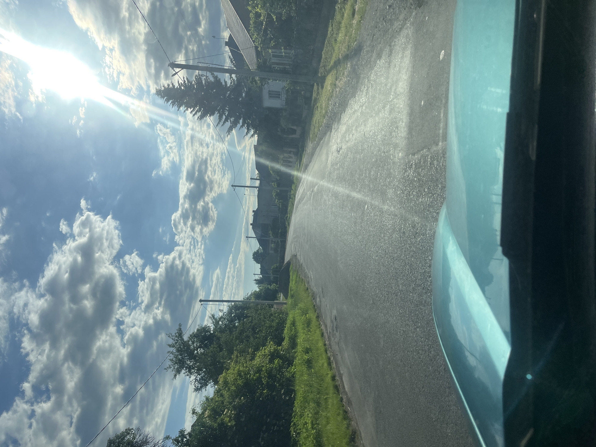 The image depicts a view from inside a vehicle looking out onto a rural road in Tarnivtsi, Ukraine. The road is paved and appears wet in some sections, with a grassy verge on the left side bordered by trees and bushes. On the right side of the road, there are houses with visible rooftops and fences, surrounded by vegetation. The sky is blue with scattered white clouds, and the sun is shining brightly, casting lens flare across the image. Utility poles with wires are visible along the roadside. The front hood of the vehicle, colored teal, is partially visible in the foreground. The scene suggests an outdoor environment under a sunny day.