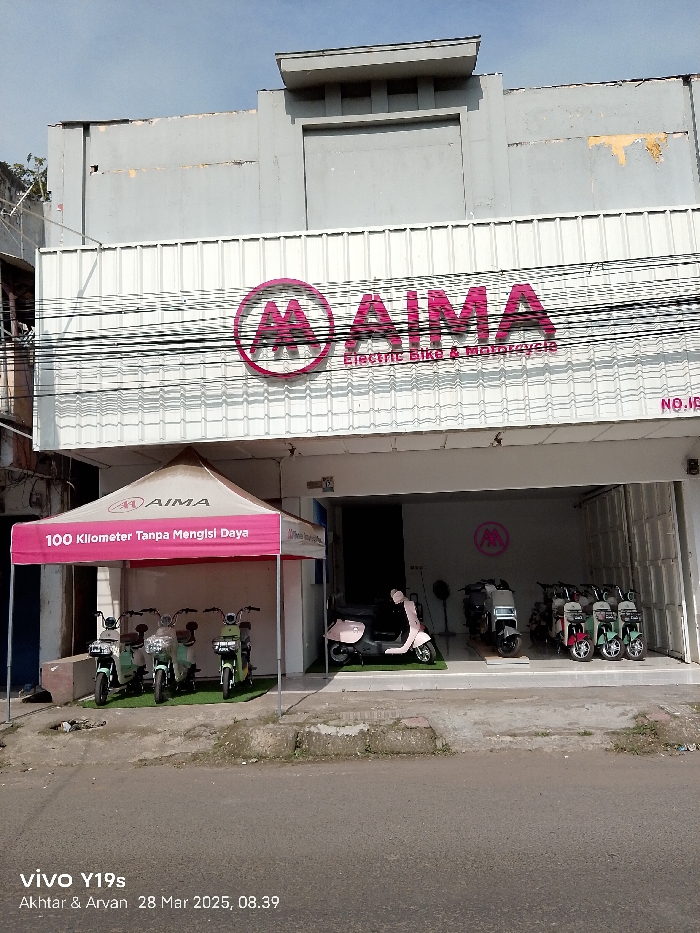 The image shows a storefront in Jatibarang, Indonesia (latitude: -6.471, longitude: 108.302). The store is called "AIMA Electric Bike & Motorcycle". A white canopy tent with pink trim advertises "100 Kilometer Tanpa Mengisi Daya" (100 kilometers without charging). The tent is in front of the store, protecting four electric motorcycles from the sun. The motorcycles are green and white. Another electric motorcycle, painted pink, is parked outside of the tent. The sign above the entrance to the store is white and has a pink "AIMA" logo with the words "Electric Bike & Motorcycle" below it. The sign also has a white "NO. 15" in the top right corner. The storefront is made of grey corrugated metal and has a large open door. The image was taken on March 28th, 2023 at 08:39.