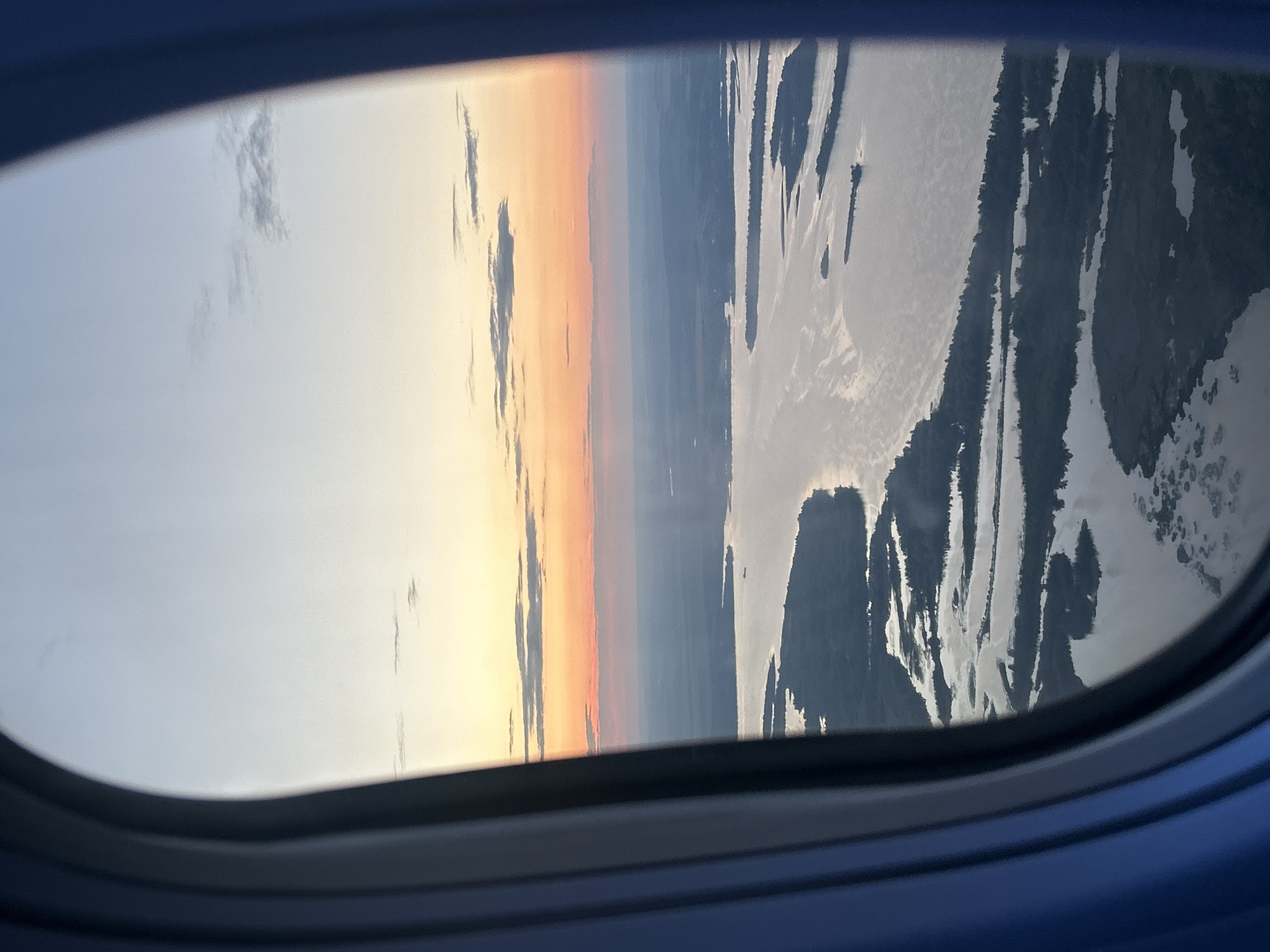 The image captures a view from an airplane window at what appears to be either sunrise or sunset. The sky transitions from a pale blue on the left to warm oranges and yellows as it moves towards the center, suggesting the sun is low on the horizon. Darker, wispy clouds are visible against the lighter sky.
The scene outside the window is a landscape seen from a high altitude. On the right side, a body of water, likely a river or lake, is prominent. Its surface reflects the sky, creating streaks of light and dark. The landmasses bordering the water are dark and appear to be covered in vegetation, possibly forests or trees, given the elongated, textured shapes. The overall impression of the landscape is rugged and natural.
The setting is clearly an airplane, indicated by the dark blue frame of the window in the foreground. The image is oriented vertically, meaning the view is seen from a side window, and the horizon is a vertical line running through the image. There are no people or specific objects clearly identifiable in the landscape, and no text is visible in the image. The time of day is indicated by the sky's colors, and the weather appears clear, with only a few clouds. The location context suggests this view is over Kokkorevo, Russia.