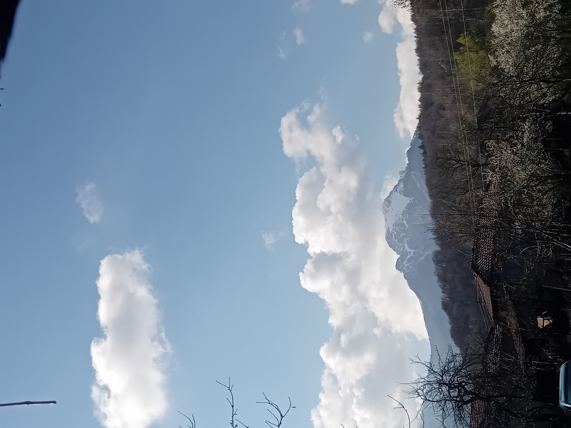 The image shows a sky with scattered white clouds, suggesting fair weather. In the background, there's a mountain range with some snow visible on the peaks. The foothills are covered with trees, some with early spring foliage and some still bare. In the lower right corner, there are hints of buildings with reddish tile roofs partially obscured by trees. The location is Unknown, Unknown.