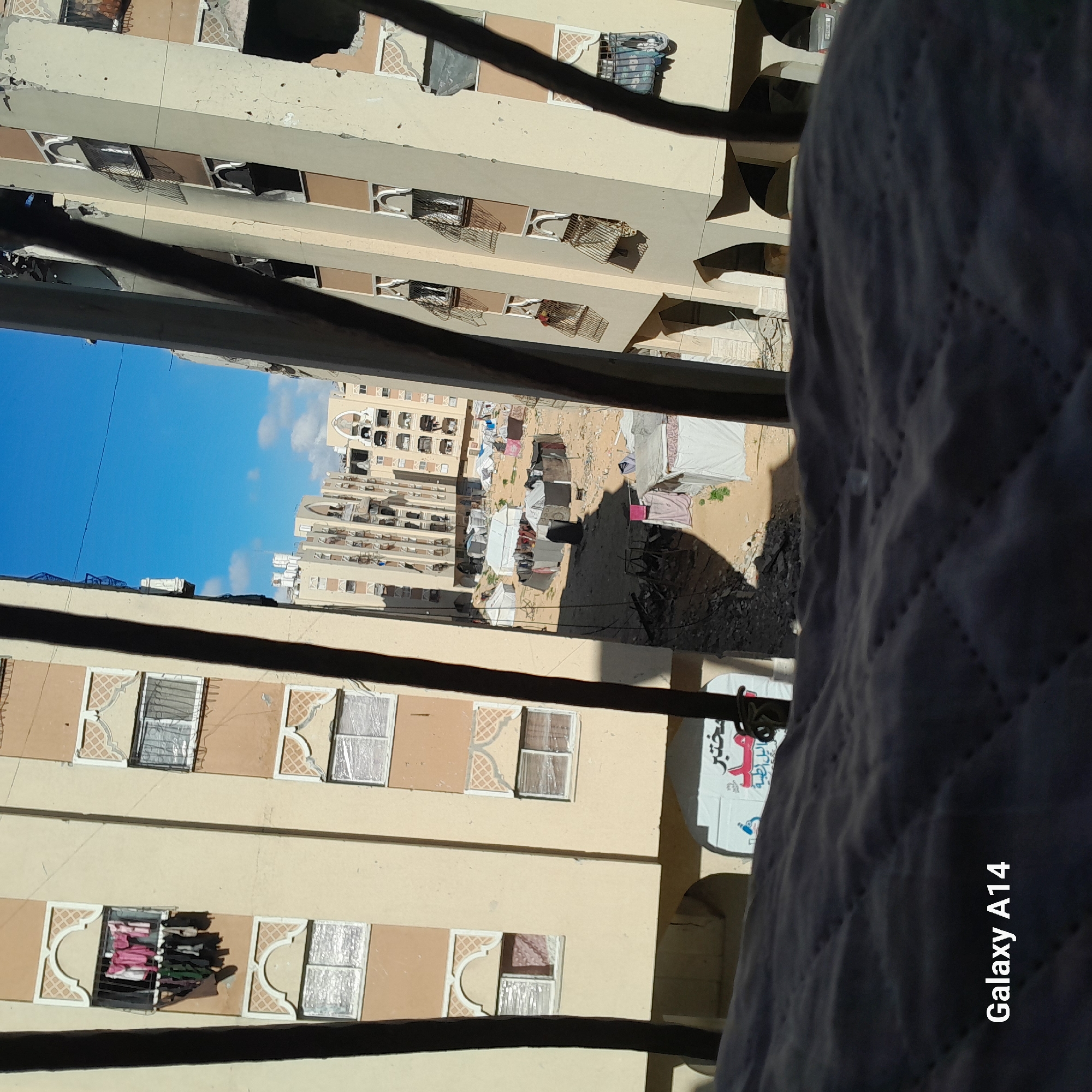 From an elevated vantage point, the image captures a view of densely built apartment buildings in an urban setting, situated in the city of Unknown, Unknown. The buildings have a light, tan-colored facade with windows that exhibit patterned or lattice-like designs. Some windows are open, revealing clothing or other items hanging out to dry. In the space between the buildings, there are makeshift structures or tents, suggesting a temporary or informal settlement. The sky above is clear and blue, with a few scattered clouds. A portion of a balcony or railing is visible in the foreground, along with what appears to be a person's draped garment, partially obscuring the view. The "Galaxy A14" watermark is visible in the lower right corner of the image.