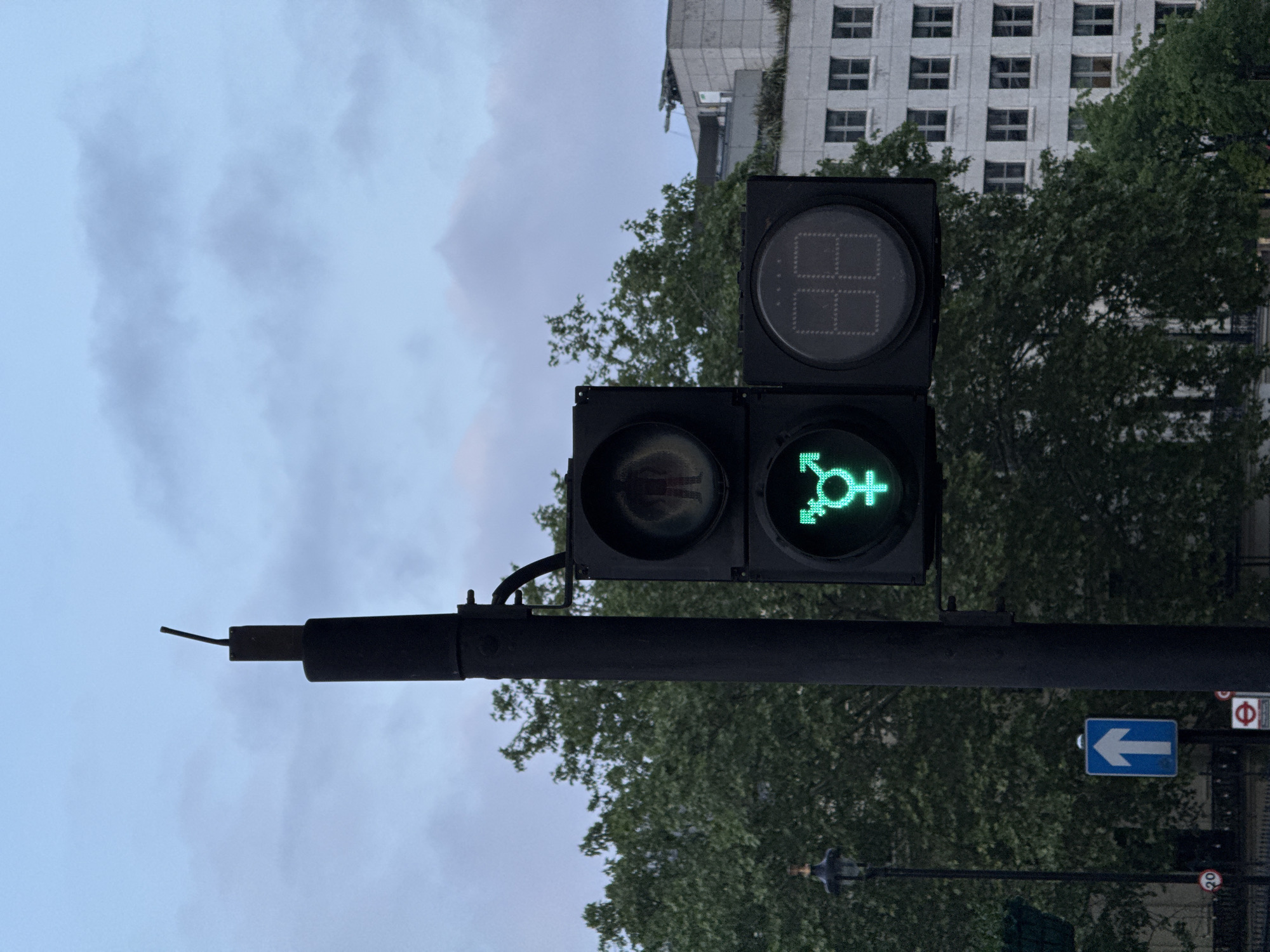 A street-level view depicts a traffic signal pole with three lights. The top light has a digital display, while the middle light appears opaque. The bottom light is green with a transgender symbol. A left turn sign is visible to the right and below the signal. Trees line the street behind the traffic signal, and a multi-story building is partially visible above the trees. The sky is cloudy. The image captures a unique traffic signal design in London, United Kingdom.