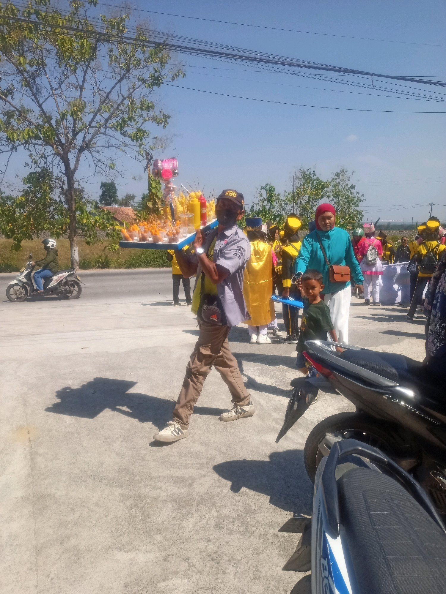 This is a medium shot photograph taken outdoors, likely on a street in Sumberlawang, Indonesia, under a clear blue sky. The foreground is dominated by a man walking from right to left, carrying a tray of food items. He is wearing a grey shirt with a black and yellow emblem on the left sleeve, tan pants, and beige sneakers. A dark bag is slung across his chest. The tray appears to hold numerous small servings of fried food, possibly corn dogs or similar snacks, with bottles of condiments like ketchup and mustard also present.
Behind the man, a group of people are walking. Several individuals are wearing yellow shirts and hats, suggesting they might be part of a group or event. A woman in a turquoise long-sleeved shirt and a red headscarf is walking towards the left, holding a brown purse. A young boy in a green t-shirt is walking beside her, holding her hand.
To the left of the frame, a person wearing a green jacket and a white helmet is riding a white and black motorcycle away from the viewer. In the background, there are trees, some with sparse foliage, and a bright blue sky with thin power lines crisscrossing it. The ground is paved with concrete. The overall impression is that of a procession or parade.