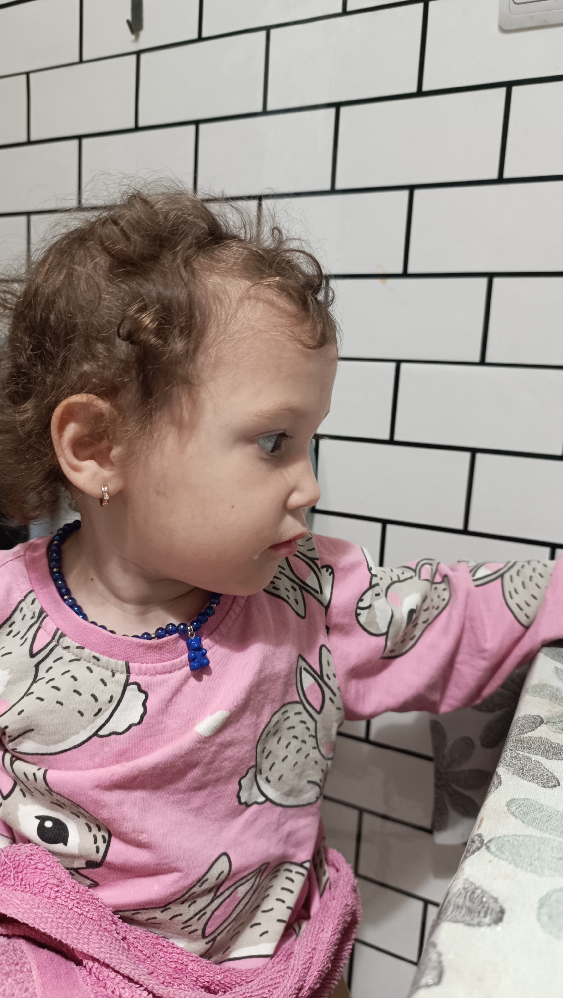 In a medium close-up shot, a young child with curly brown hair and light skin is pictured from the chest up, facing to the right of the frame. The child is wearing a pink long-sleeved shirt adorned with a repeating pattern of cartoon rabbits. A dark blue beaded necklace with a blue bear-shaped charm is visible around the child's neck. A small gold hoop earring is present in the child's left ear. A pink towel is draped over the child's lap. The background consists of a white subway tile wall with dark grout lines. The scene appears to be indoors, possibly in a home setting in Chaikovsky, Russia.