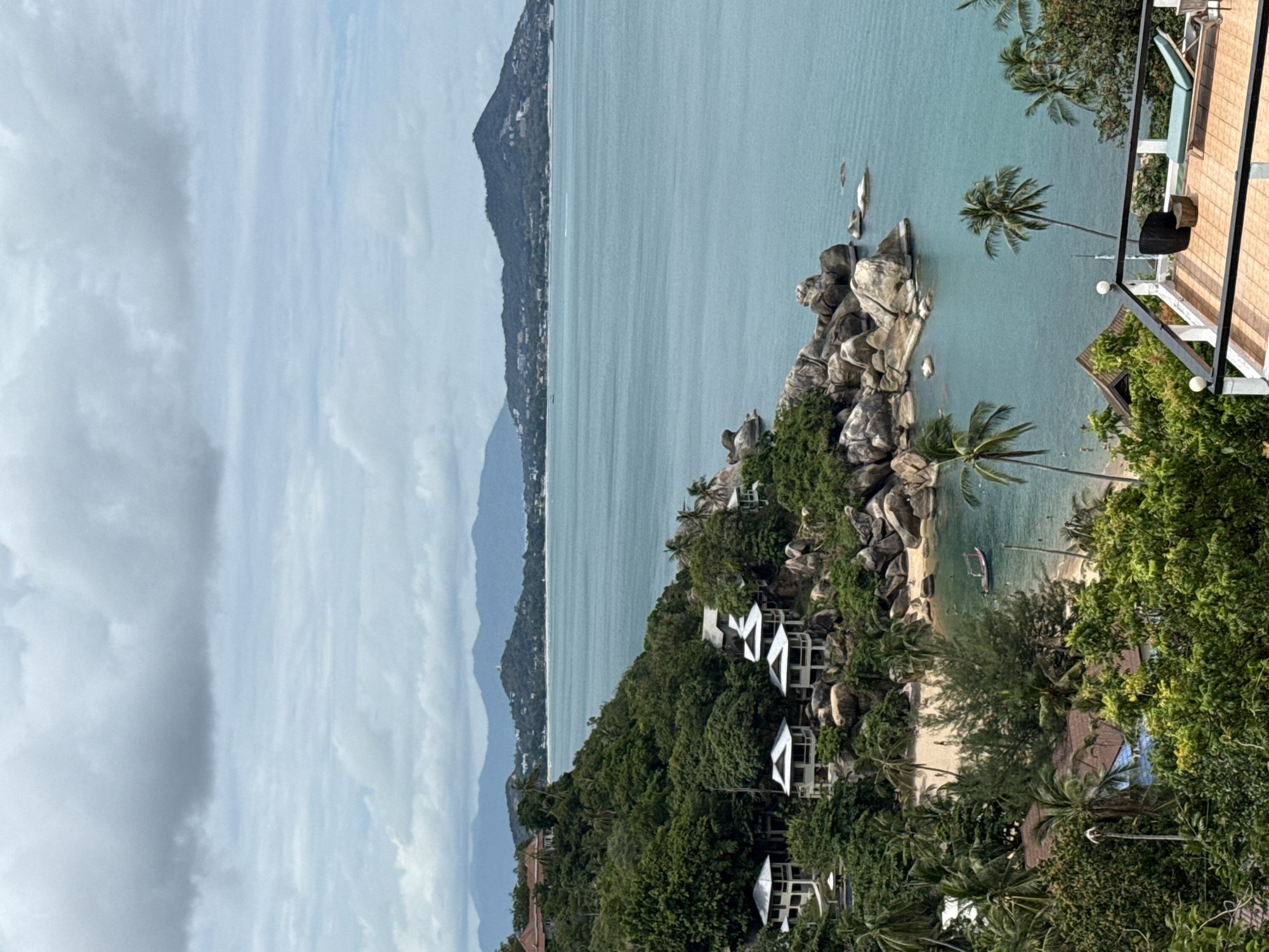 This image captures a scenic view of a tropical coastline in the Koh Samui District of Thailand. In the foreground, a tiled balcony with white railings features a lounge chair, a small table, and a black wicker bin. Beyond the balcony, lush green trees and palm trees dominate the landscape, interspersed with several white bungalows with grey roofs nestled on a rocky incline. A small, sandy cove with calm, turquoise water is visible below, with a small red and white boat anchored in the bay and a few people wading near the shore. The water extends to the horizon, where a range of hazy, green hills form the backdrop. The sky is overcast with soft, grey clouds. The scene suggests a peaceful, resort-like environment.
